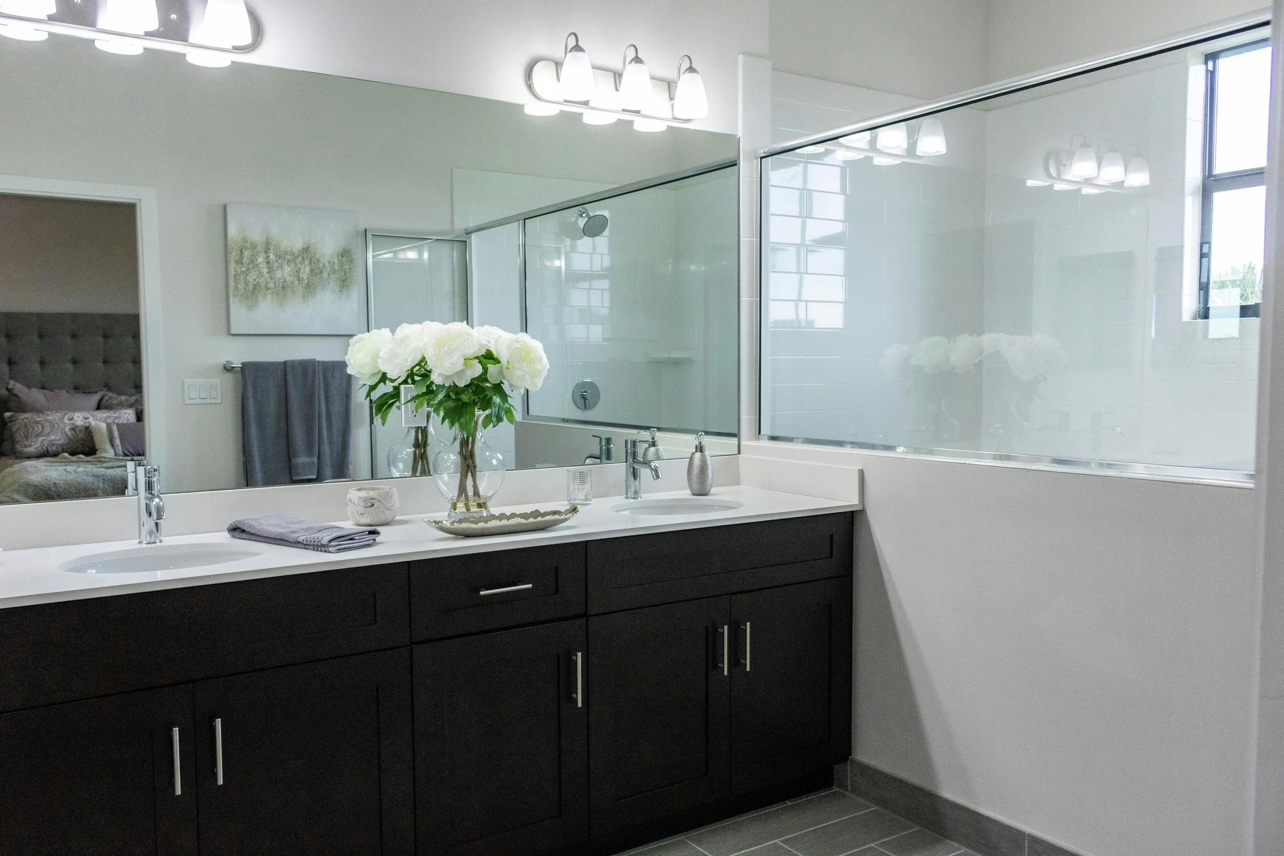 Modern bathroom with double sink vanity, large mirror, flower arrangement, glass shower enclosure, and natural light from window.