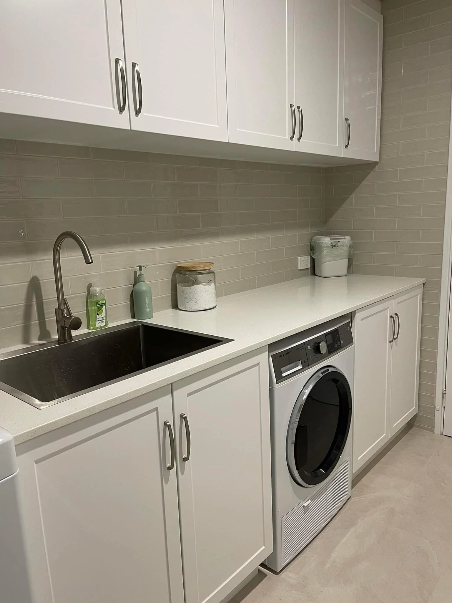 Kitchen laundry area with white cabinets, a black sink, a washing machine, and various cleaning supplies on the countertop.