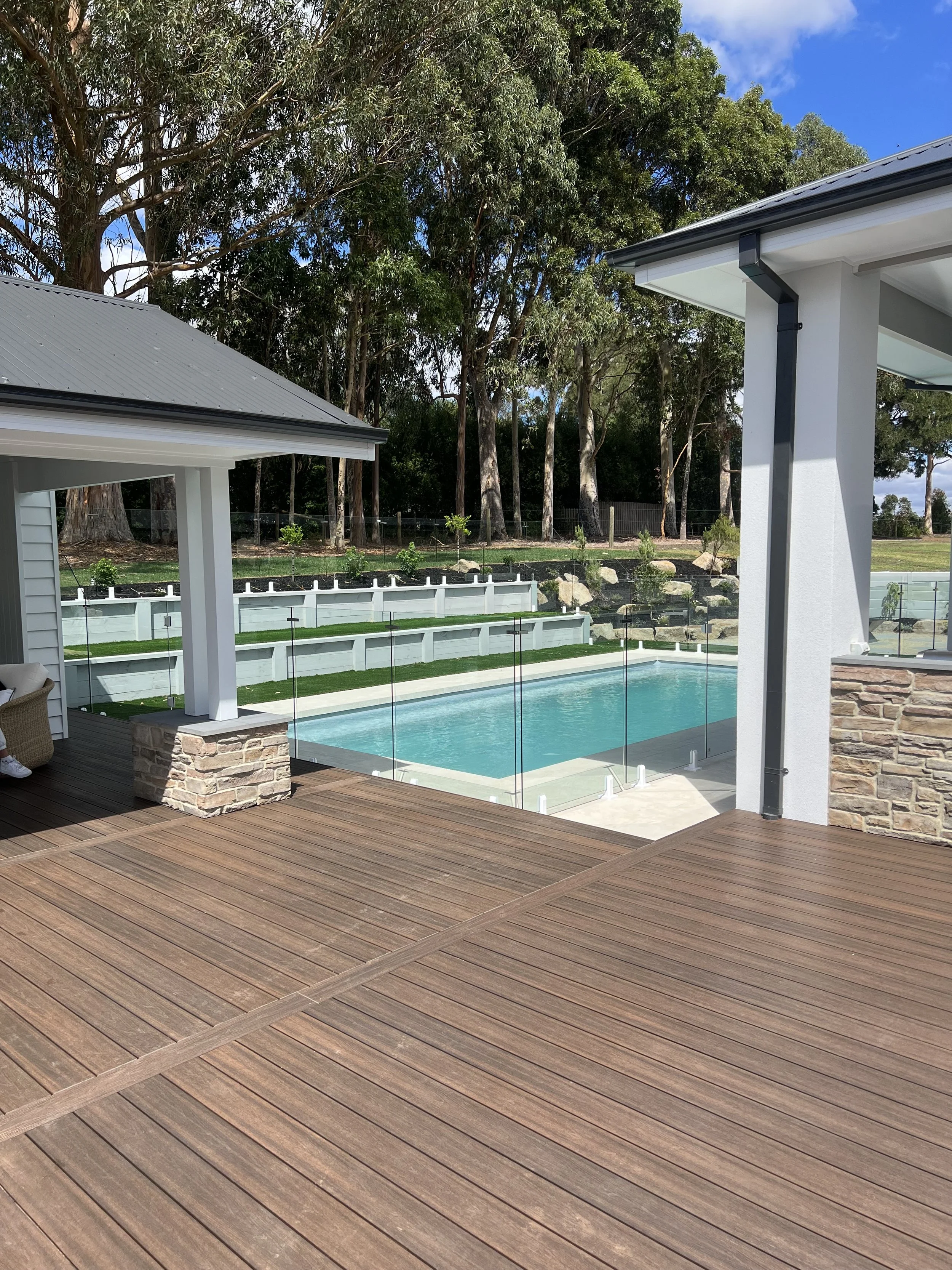 View of a backyard with a swimming pool surrounded by a wooden deck, glass fencing, and green lawn. Tall trees and blue sky in the background.