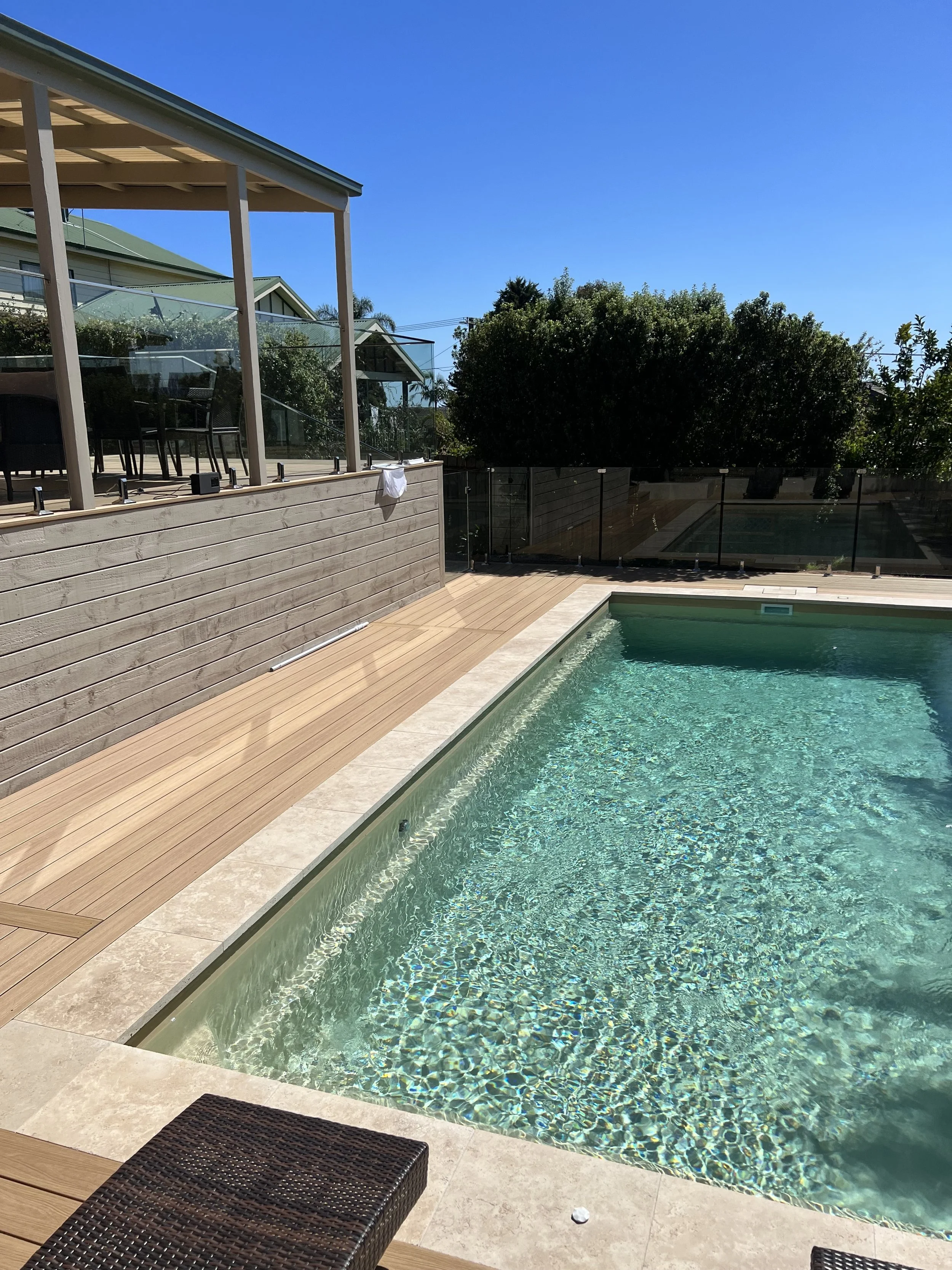 A backyard swimming pool with clear water, surrounded by a wooden deck and a beige tiled border. There is a wicker lounge chair in the foreground and a covered patio with black chairs and a glass railing in the background. Green bushes and trees are visible beyond the pool area, under a bright blue sky.