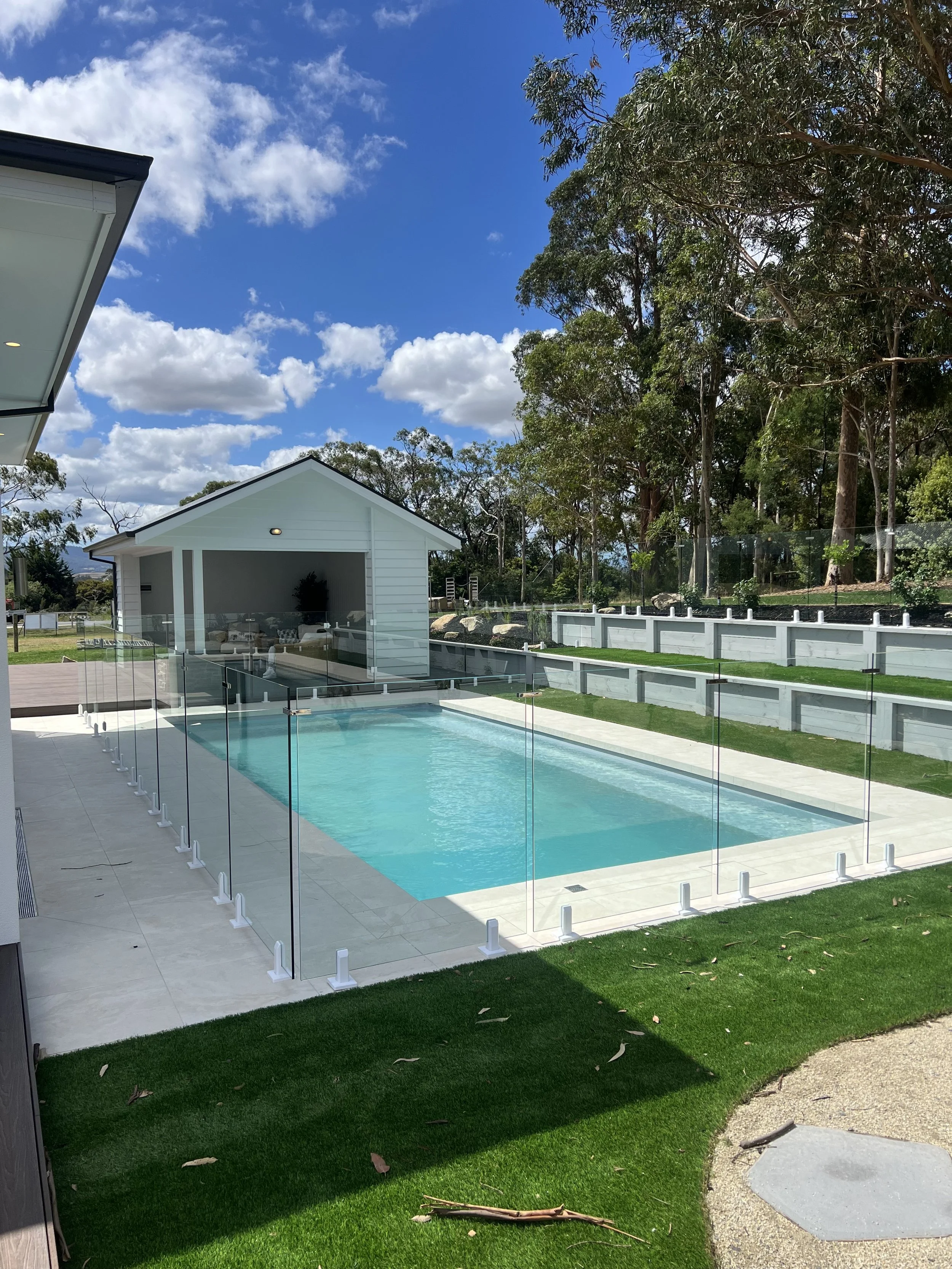 A modern backyard with a rectangular swimming pool enclosed by a glass fence, a small white pool house at the far end, and a lush green lawn surrounding the area, with tall trees in the background under a blue sky with some clouds.