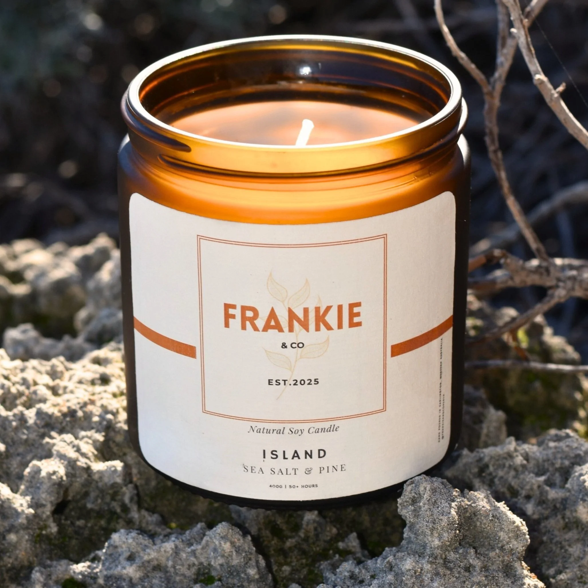 A lit natural soy candle in a brown jar with a white label, placed on rocky terrain outdoors, with twigs and shadows in the background.