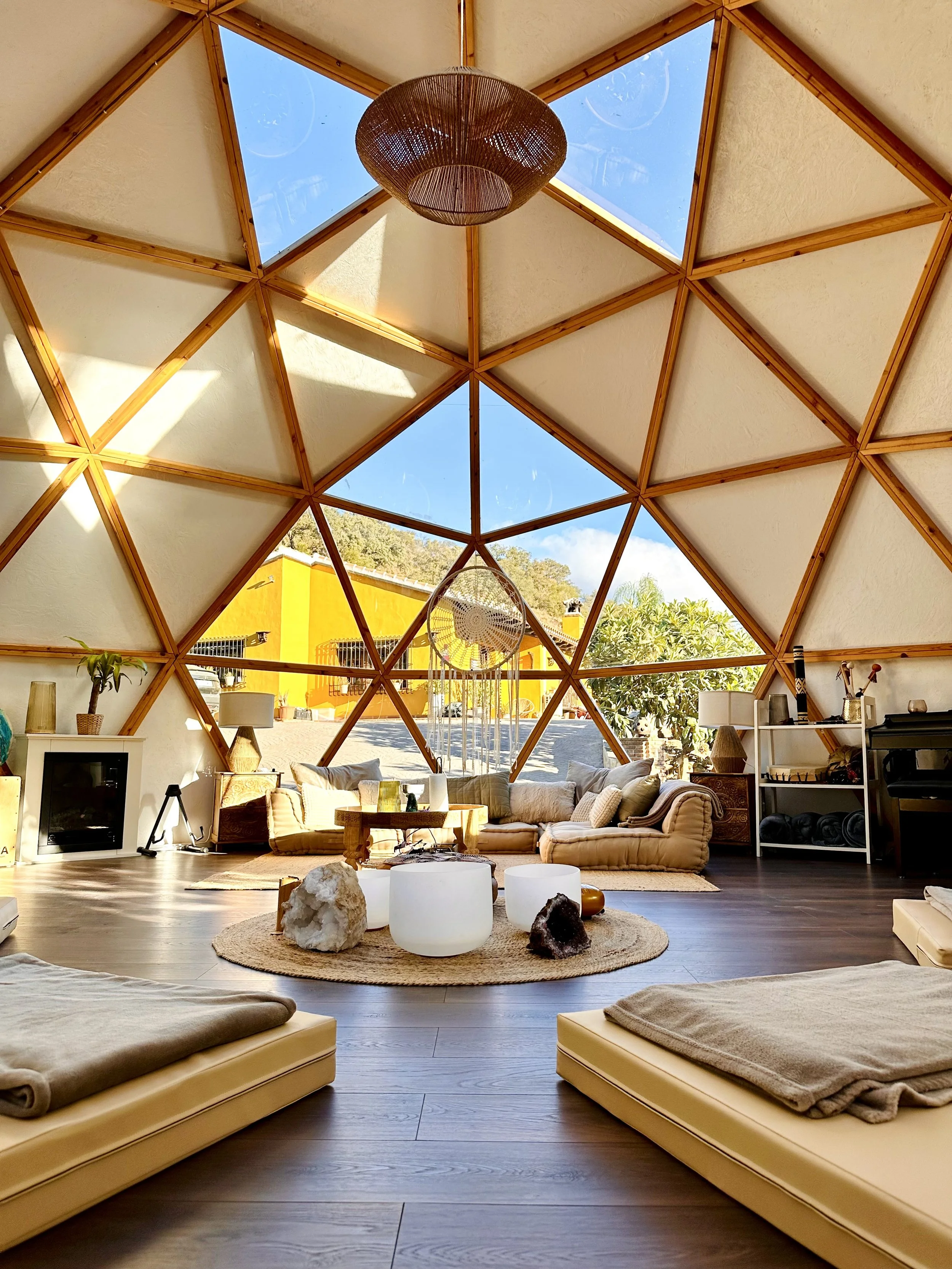 Interior of a geodesic dome with a vaulted ceiling featuring large triangular windows, sunlit living area with beige cushions and a round woven rug, decorated with natural elements like rocks and a dreamcatcher hanging from the ceiling.