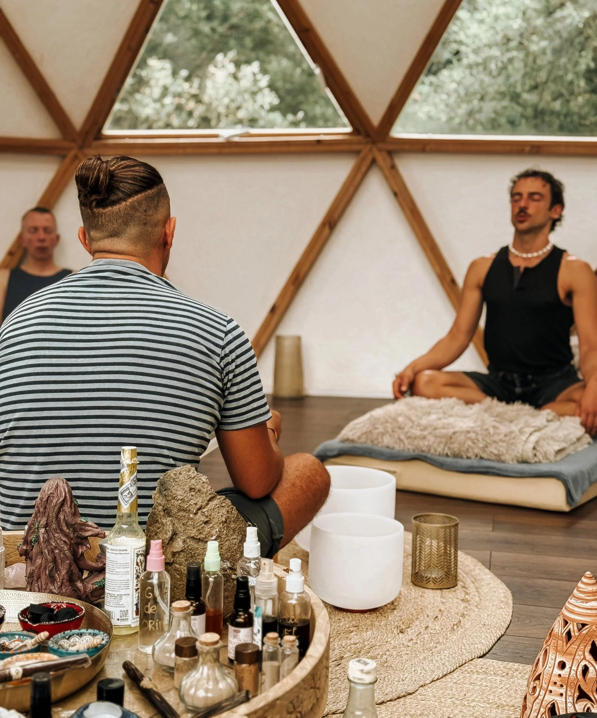 People participating in a meditation or yoga session inside a geometric-shaped wooden structure with large windows, with one person leading and others sitting cross-legged on cushions.