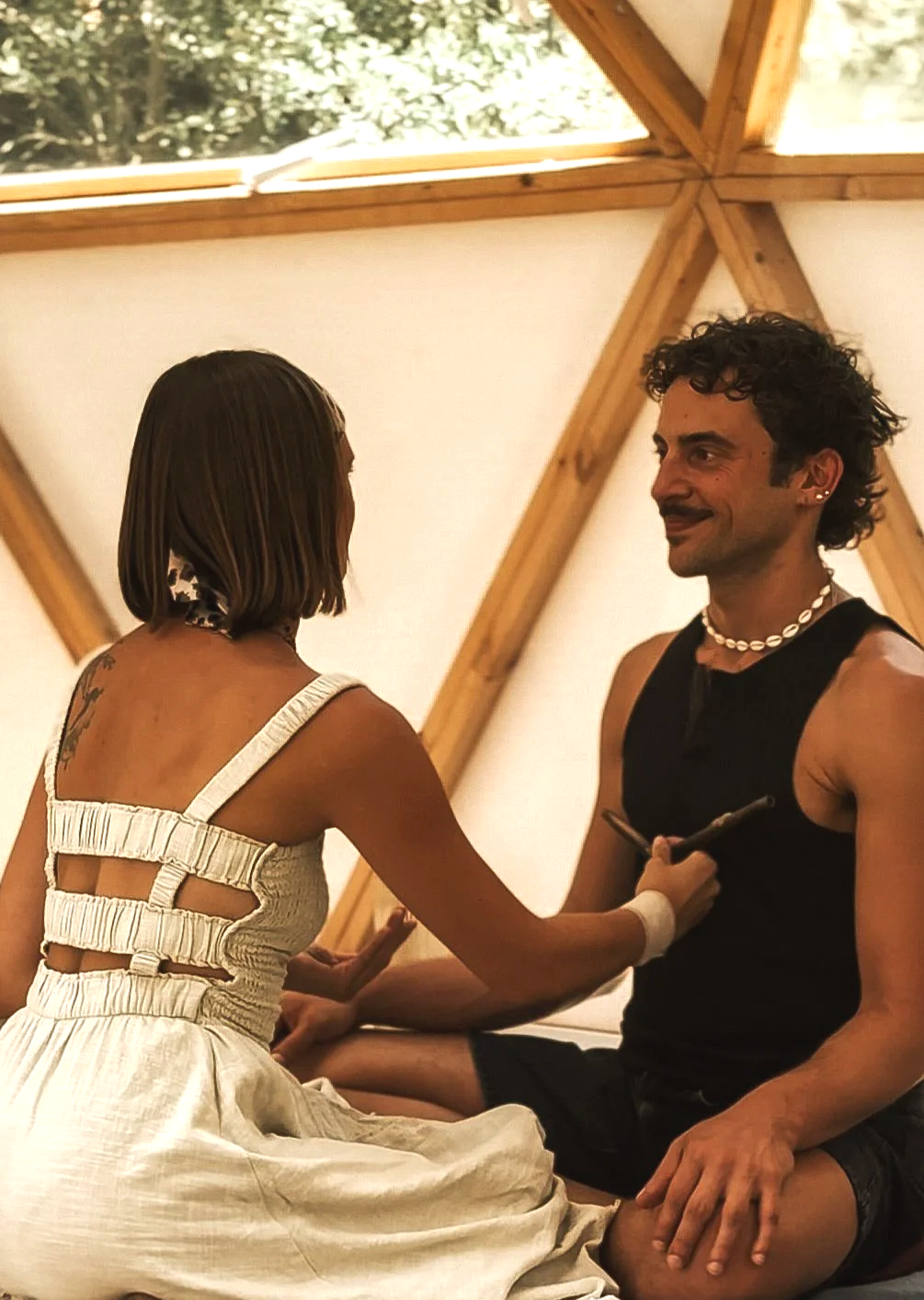 Two people sitting on the floor inside a wooden A-frame cabin, facing each other and engaging in conversation, with a skylight and trees visible through the skylight window.