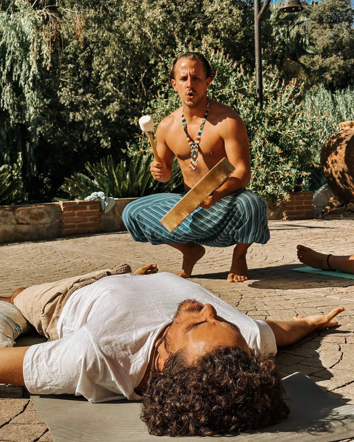 A shirtless man wearing a colorful beaded necklace and striped loose pants squats and plays a percussion instrument with a mallet while a man with curly hair and a beard lies on the ground in front of him, outdoors in a sunny garden.