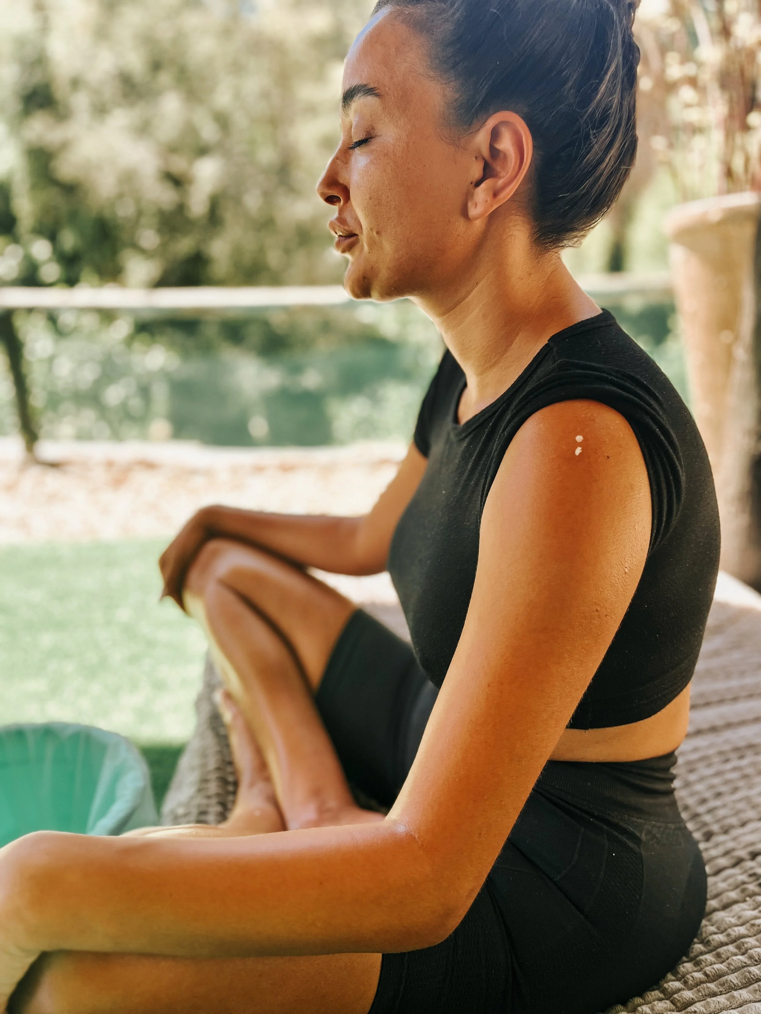 A woman in black athletic wear relaxing outdoors with eyes closed and a serene expression, sitting on a couch or outdoor furniture with a blurred background of trees and water.