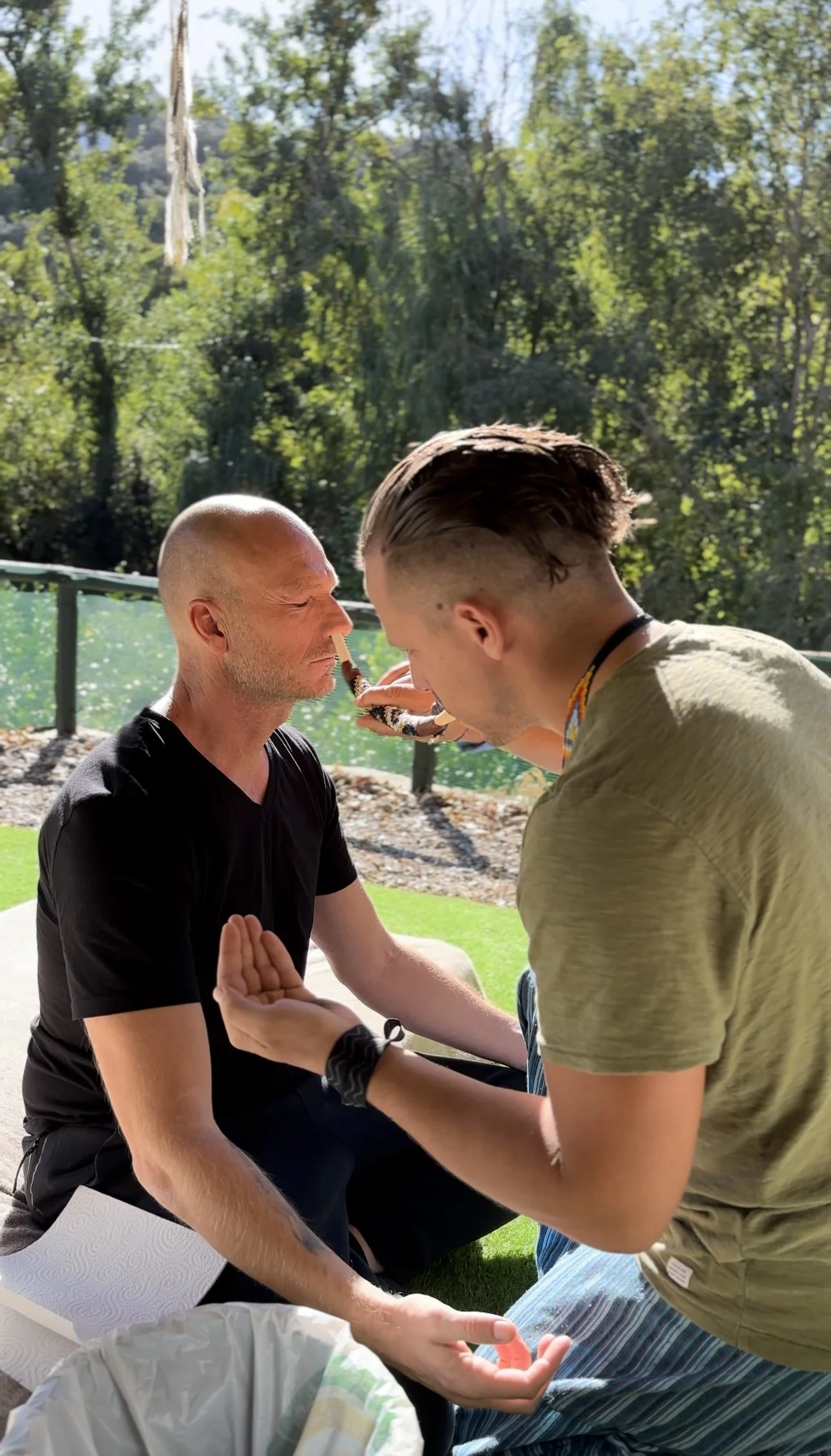Two men practicing meditation outdoors near a lake with trees in the background.