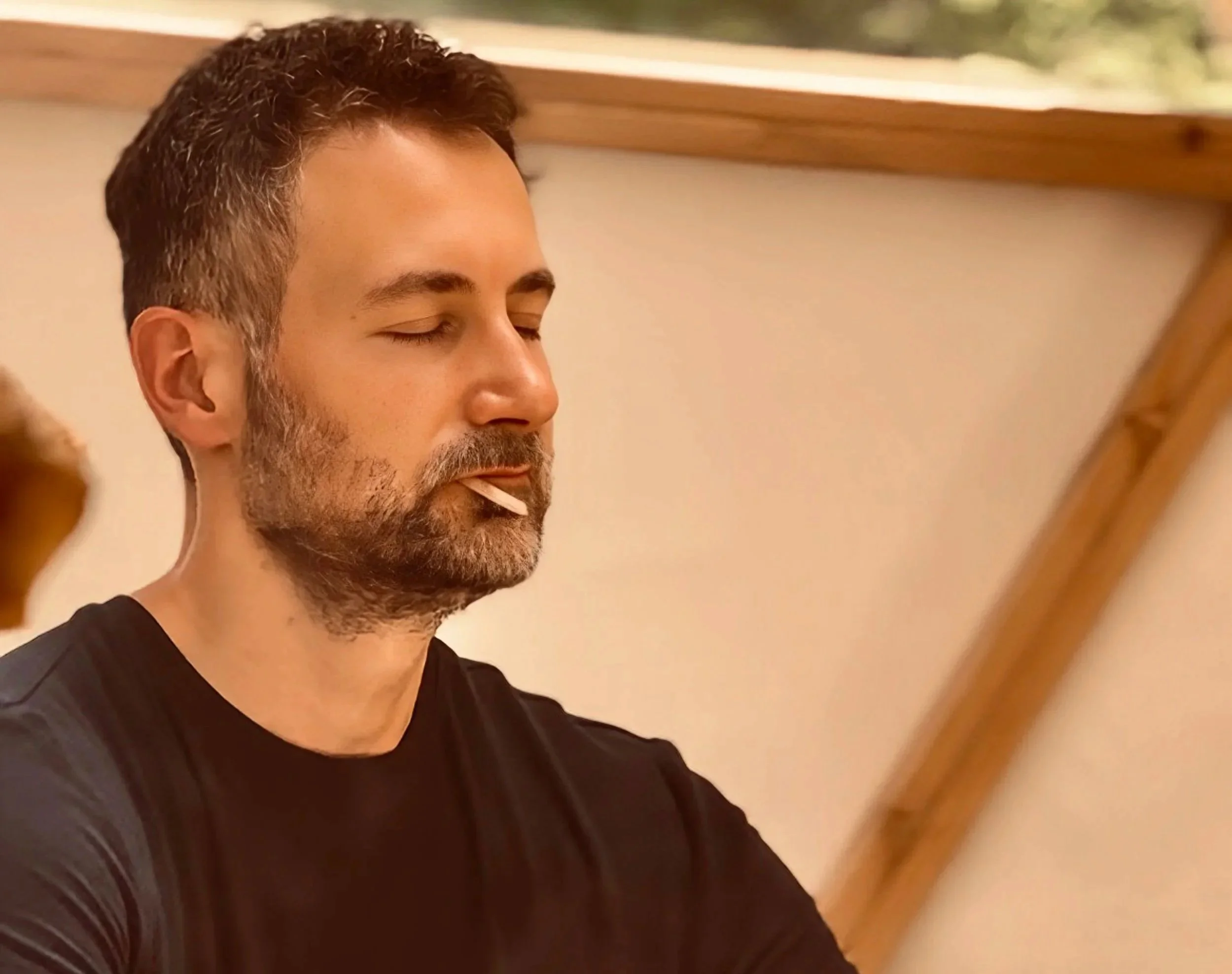 A man with short brown hair and a beard is with his eyes closed, holding a toothpick in his mouth, wearing a black shirt, inside a room with wooden beams on the ceiling.