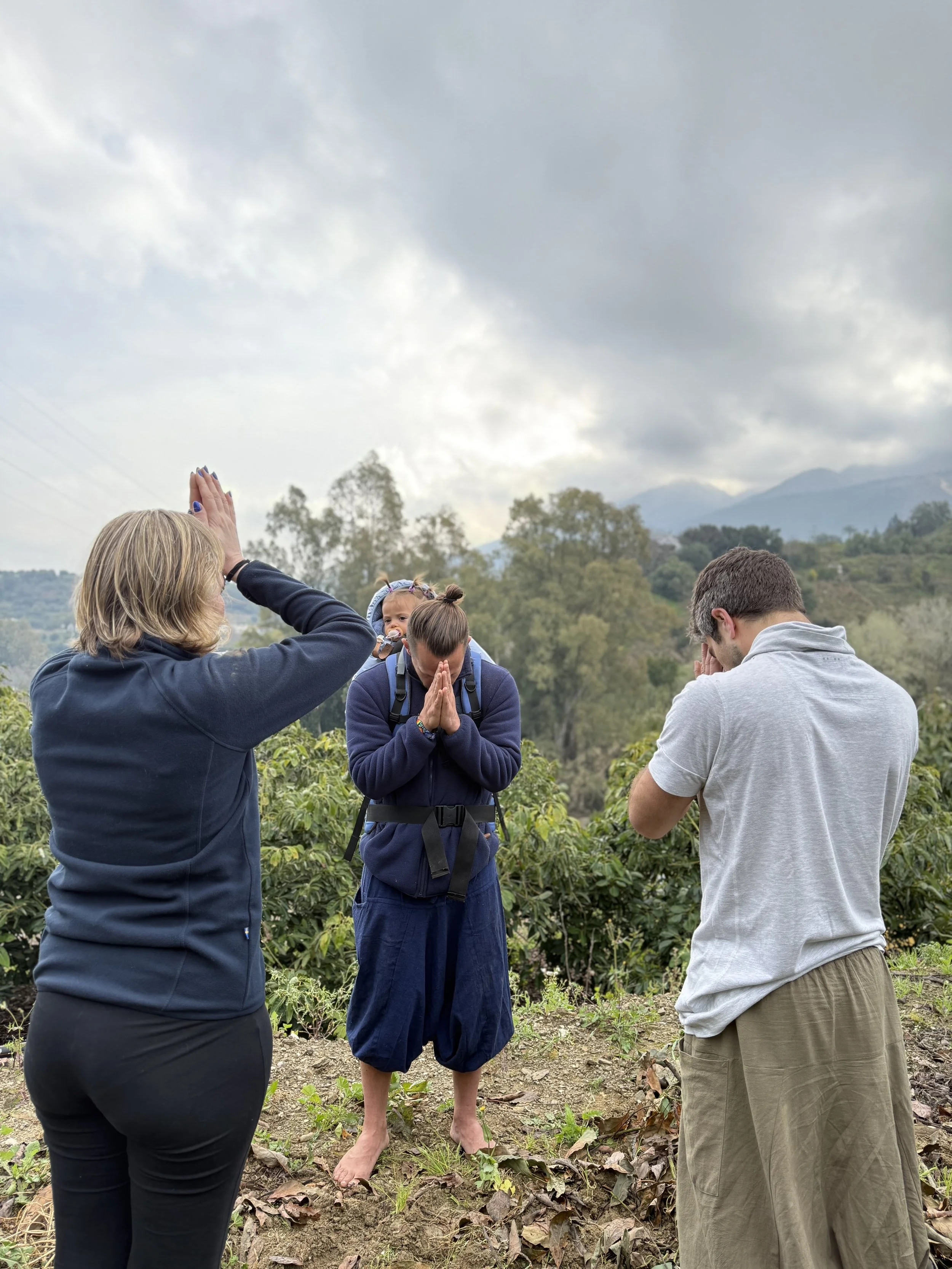 Three people are standing outdoors with lush green trees and mountains in the background. Two women and one man are seen with their hands in prayer or meditation posture. The woman on the left is raising her hand towards a woman in the center, who has a backpack and is barefoot, with her eyes closed and hands together in prayer. The man on the right has his head bowed with his hand over his face.