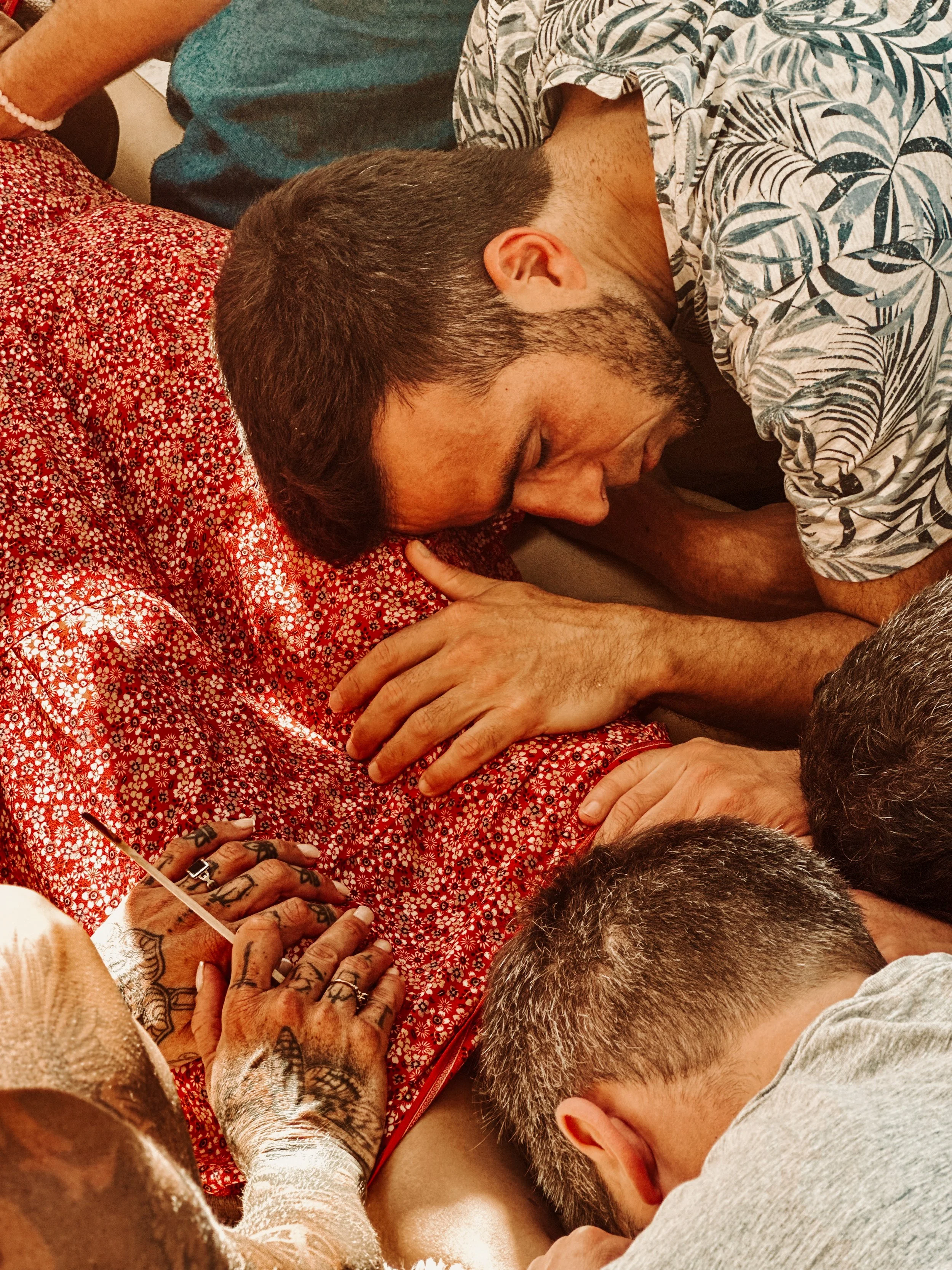 A group of people peacefully gathered in a close embrace, with one person receiving a tattoo as others hold and support them.