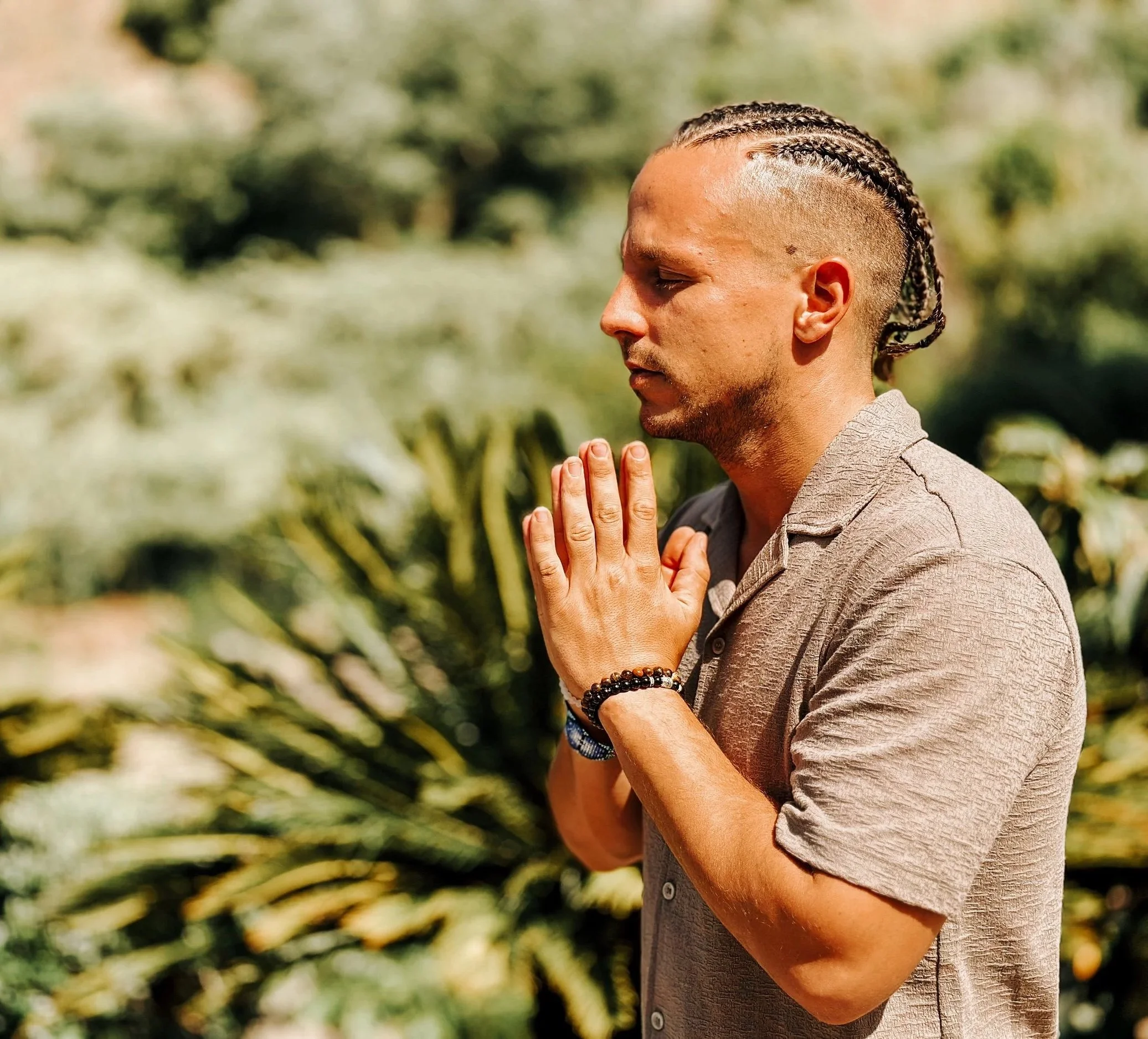A man with braided hair and a beard practicing meditation outdoors, with hands pressed together in prayer and eyes closed, surrounded by green foliage.