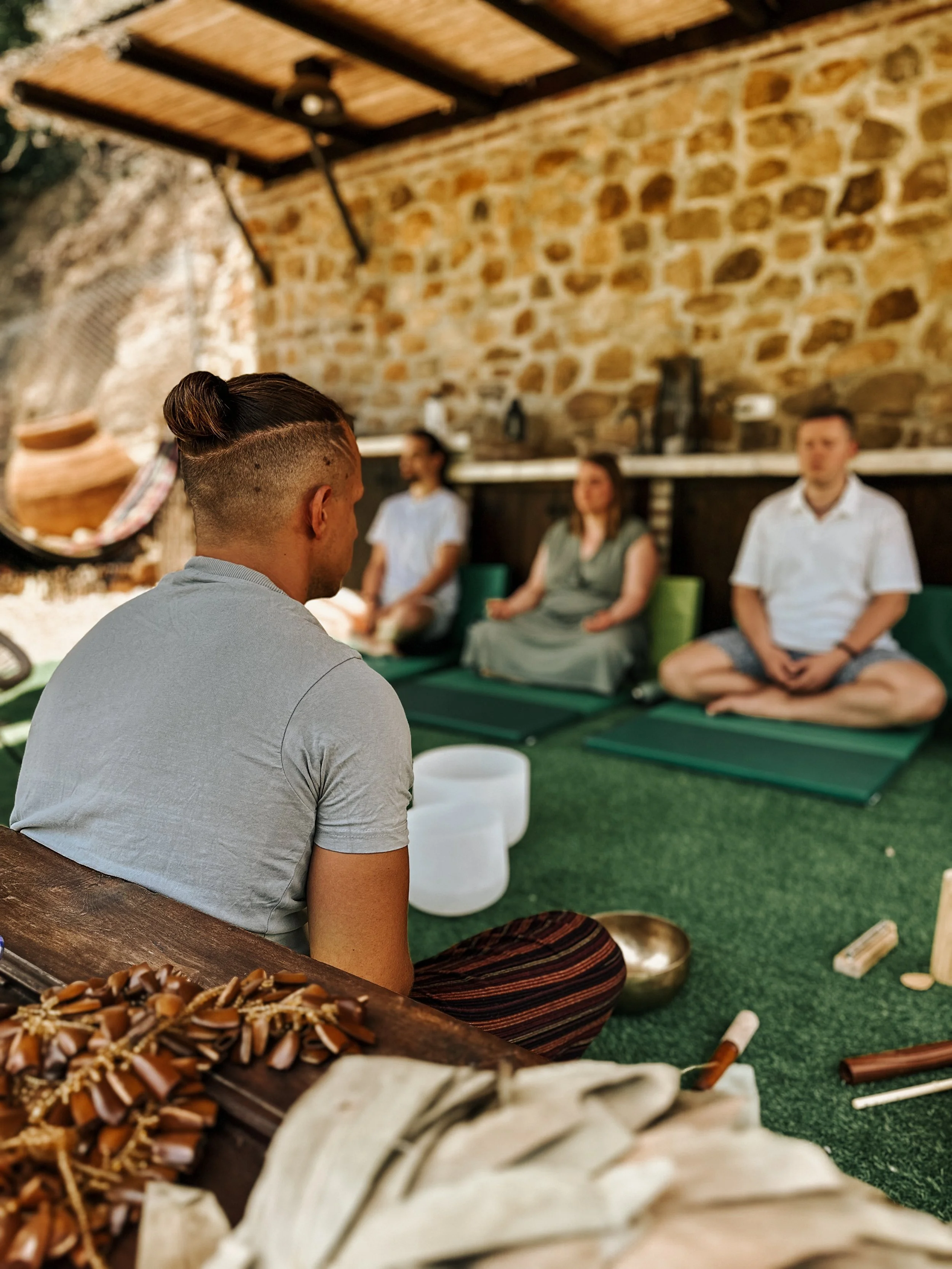 A person with a man bun seated on the ground facing a seated group of three adults on yoga mats, in a rustic outdoor setting with a brick wall background, participating in a meditation session.