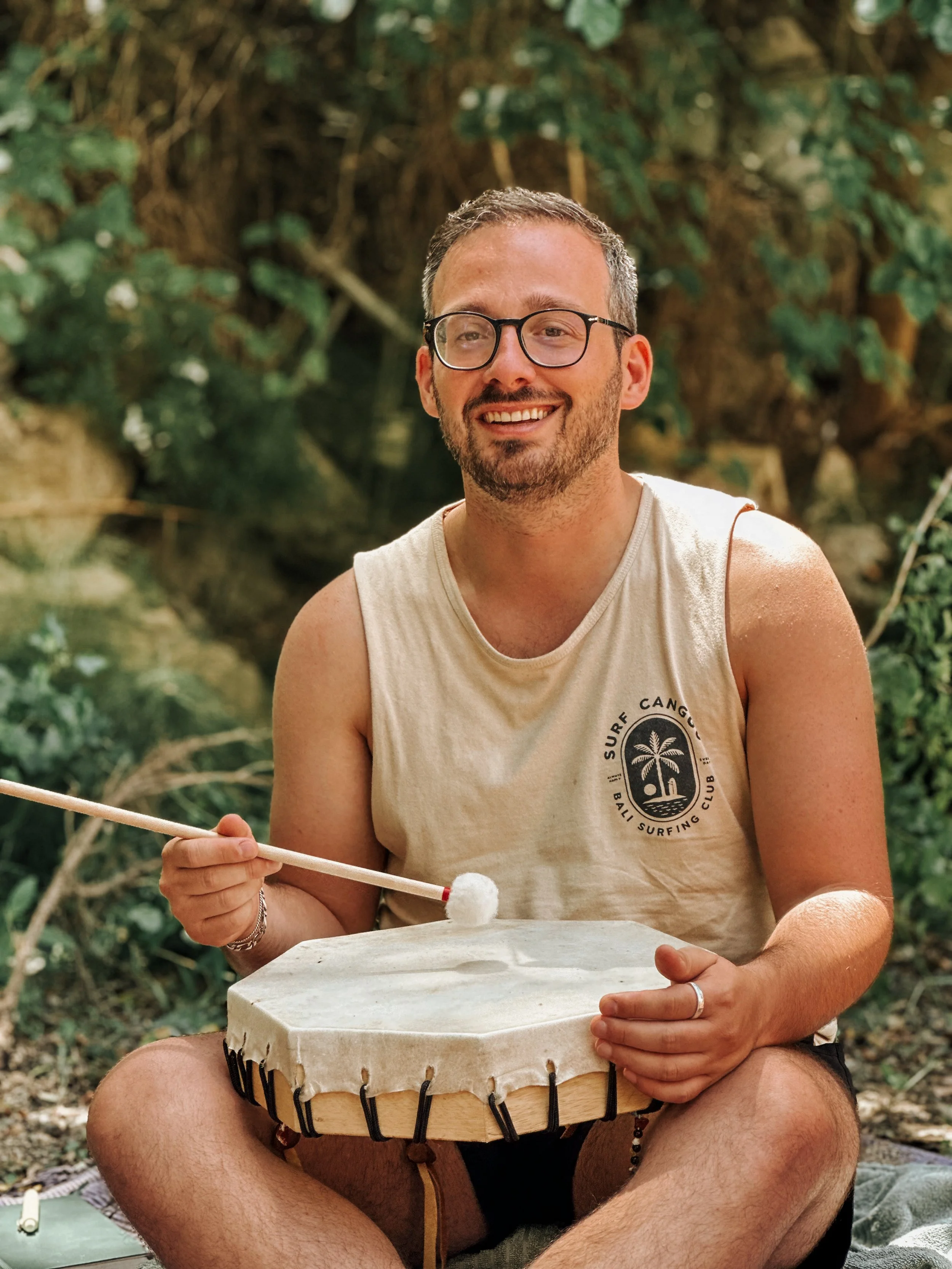 A young man with glasses, a beard, and short hair sitting outdoors in a natural setting, playing a hand drum with a stick, smiling.