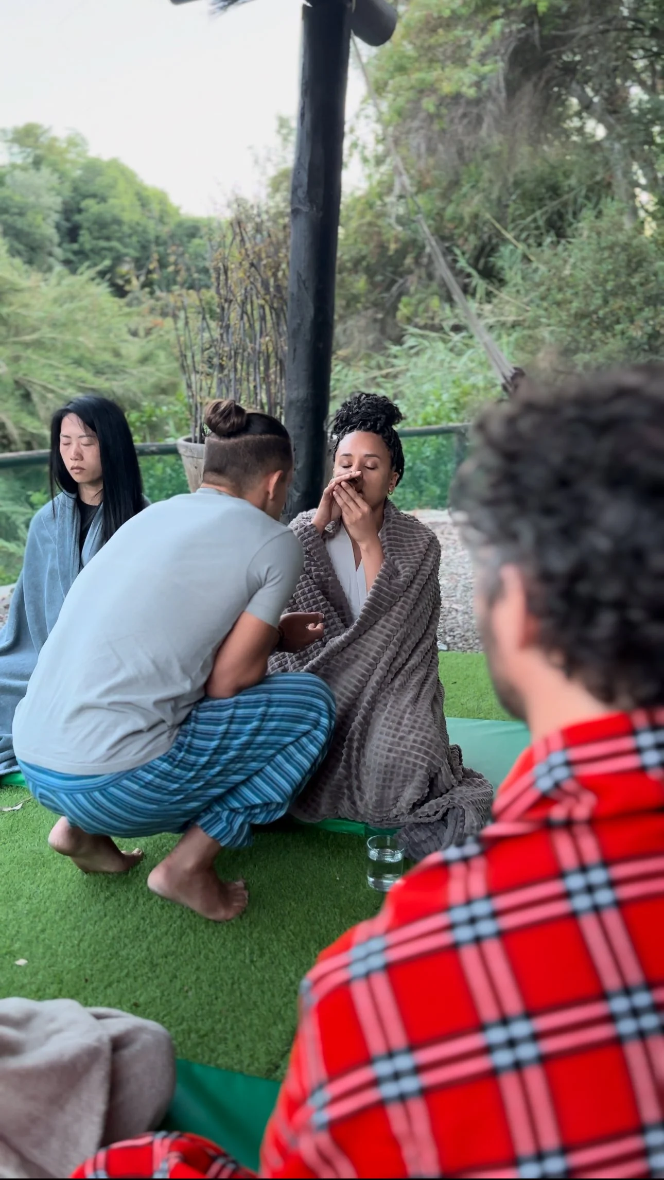 A group of people participating in a spiritual or religious ceremony outdoors. One woman appears to be praying with her eyes closed, and another person is holding her hand near her mouth. They are seated on green mats on a grassy area surrounded by t