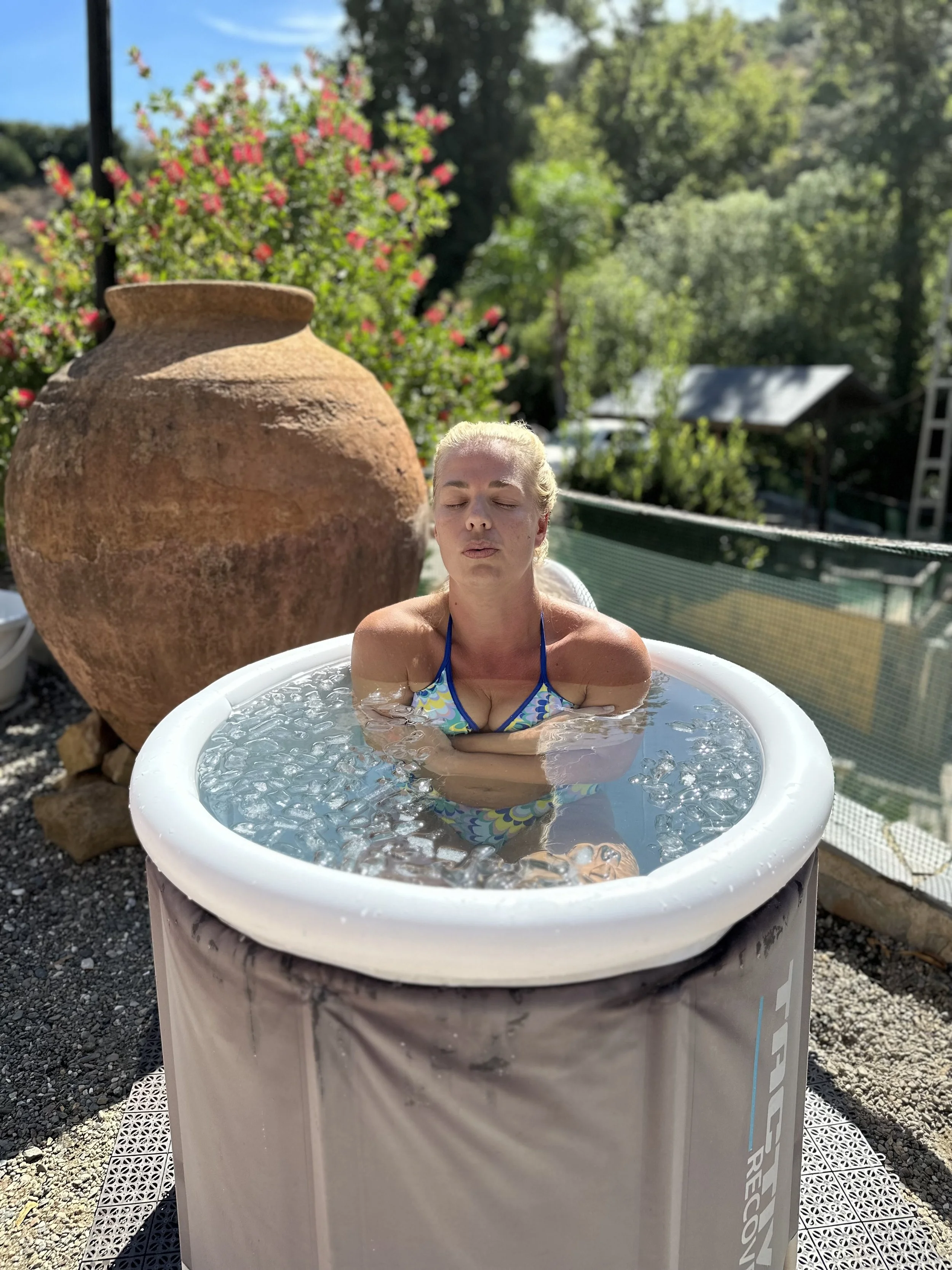 A woman with blonde hair relaxing in a portable hot tub outdoors, with a large clay pot and a lush garden with trees and pink flowers in the background.
