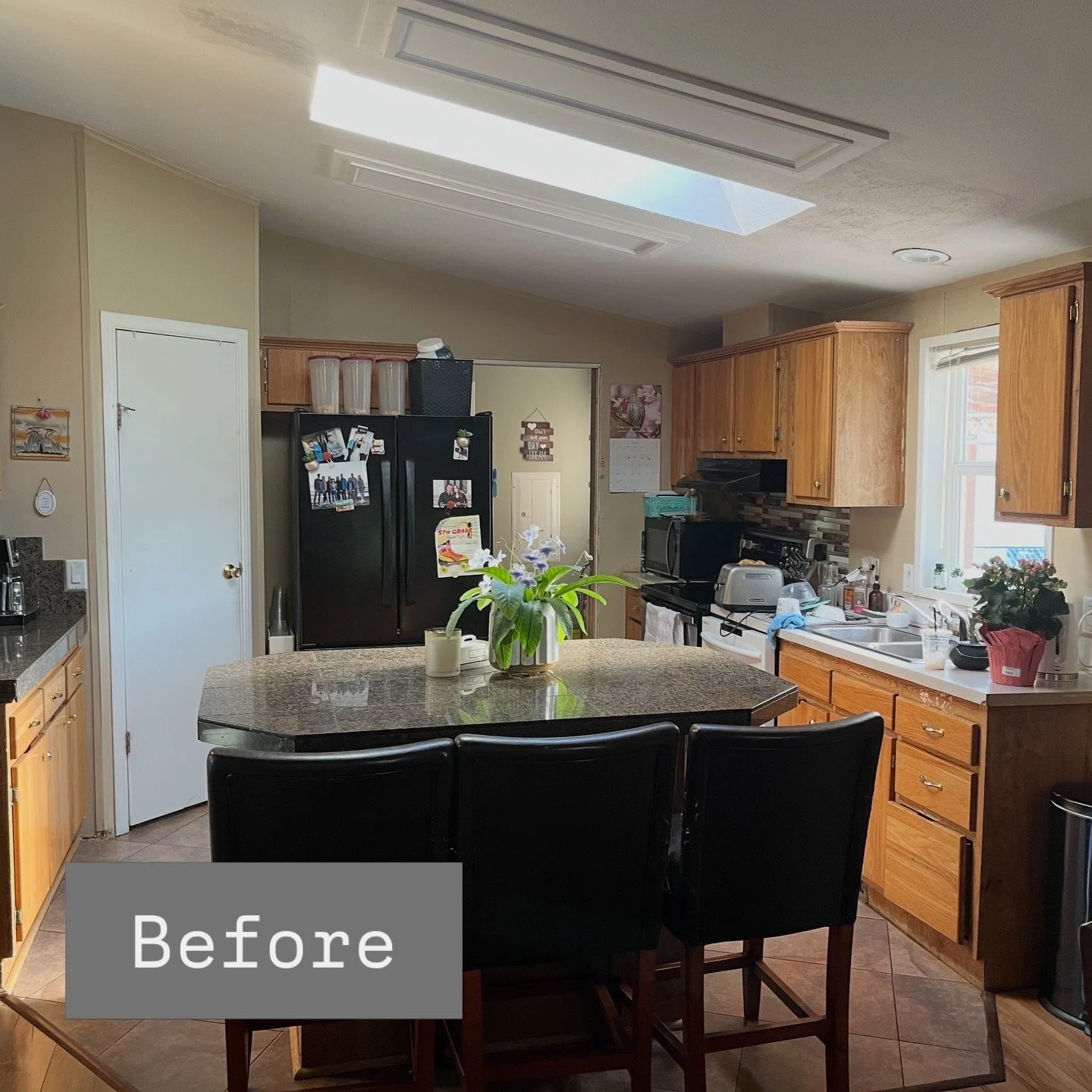 A kitchen with wooden cabinets, a black refrigerator covered with photos, a black stove, a microwave, a toaster, a sink, and a countertop with various items. There is a dining table with black chairs in the center, and a vase with flowers on the tabl
