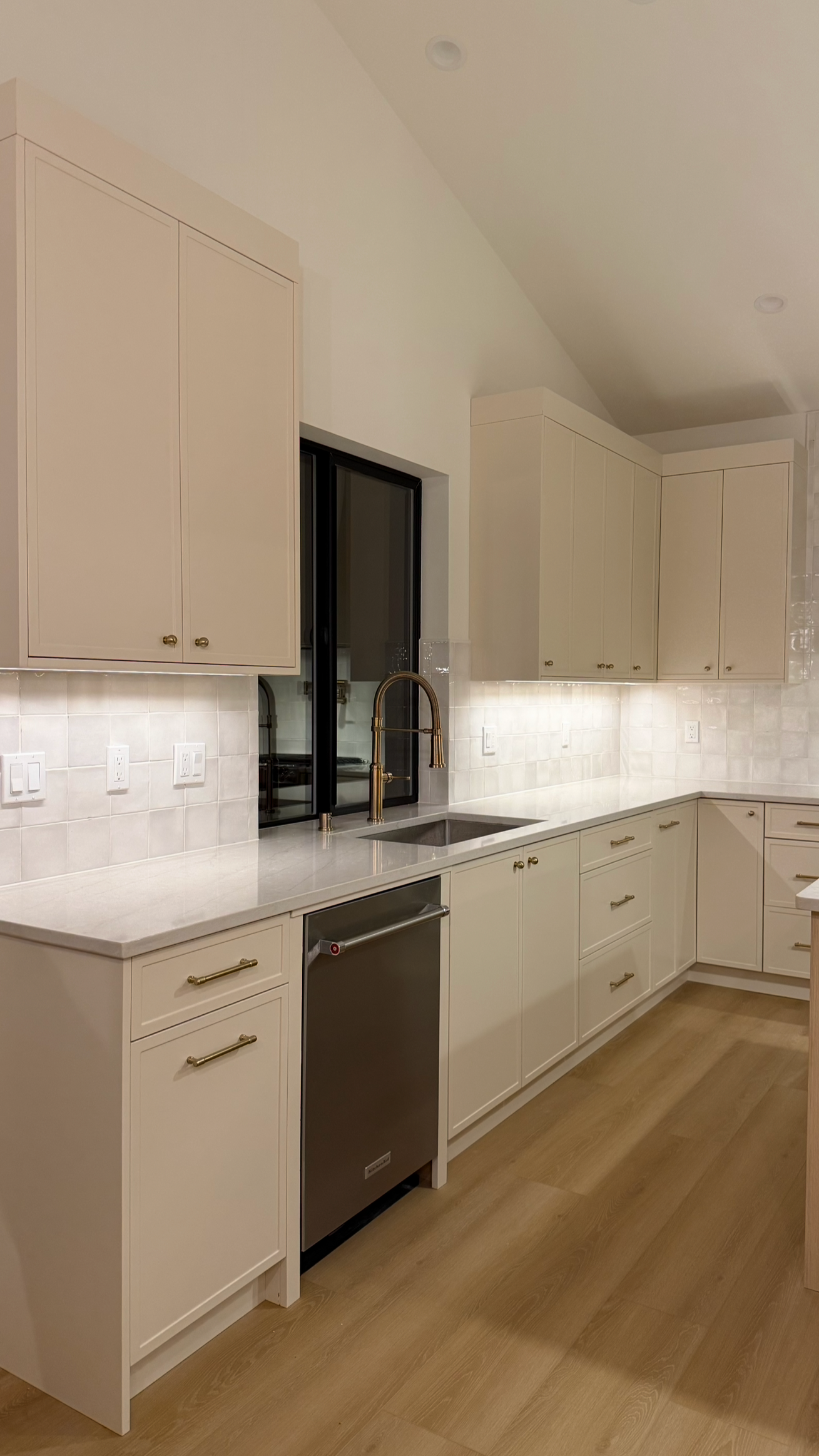 Modern kitchen with cream-colored cabinets, brass hardware, a stainless steel dishwasher, and a black-framed window above the sink with a gold faucet.