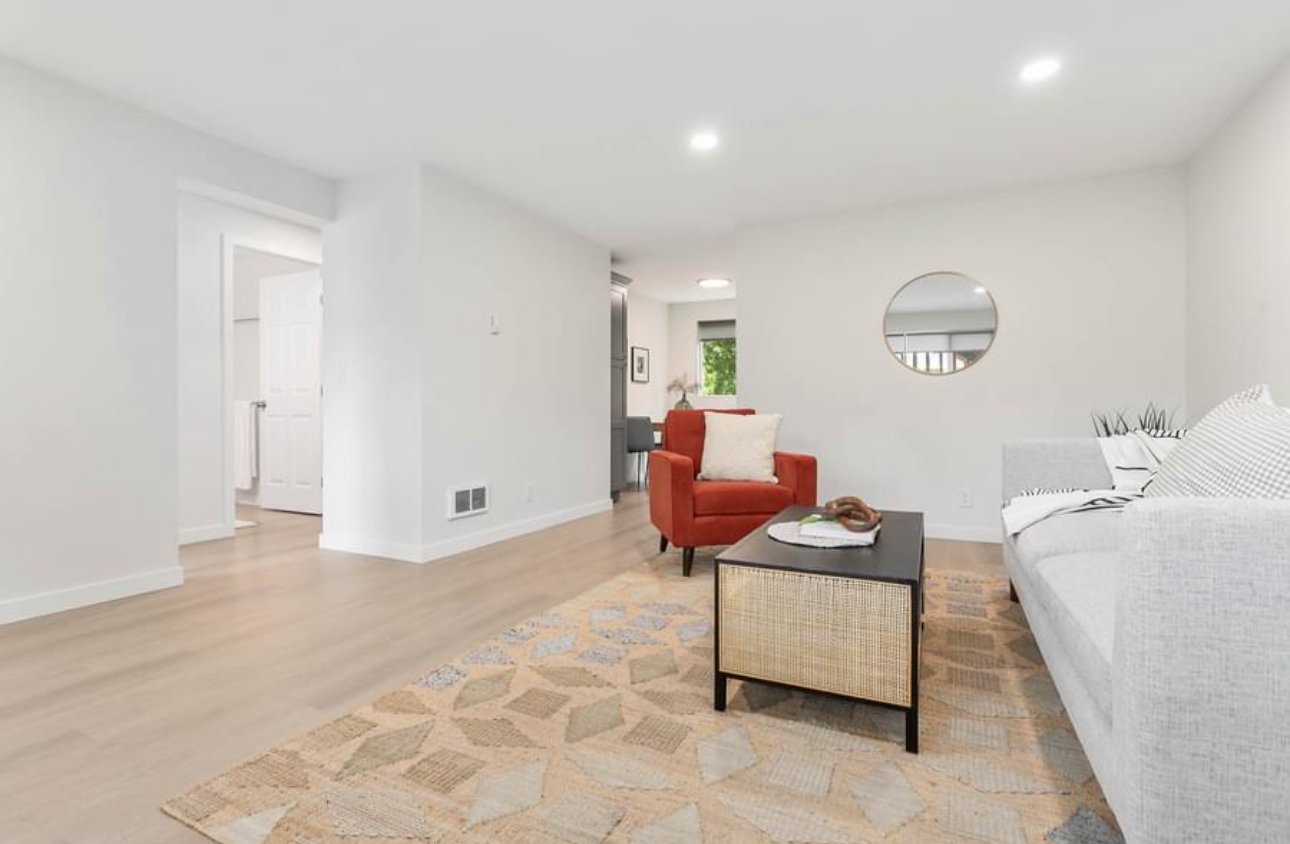 Living room with white walls, hardwood flooring, a white sofa, a red armchair with a pillow, a black coffee table with decorative objects, a patterned area rug, and a round wall mirror.