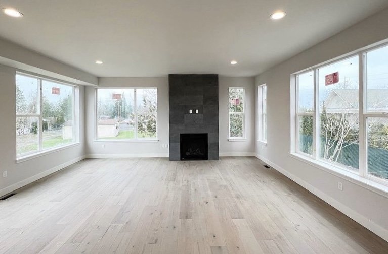 Empty living room with light wood flooring, large windows, and a central black fireplace.