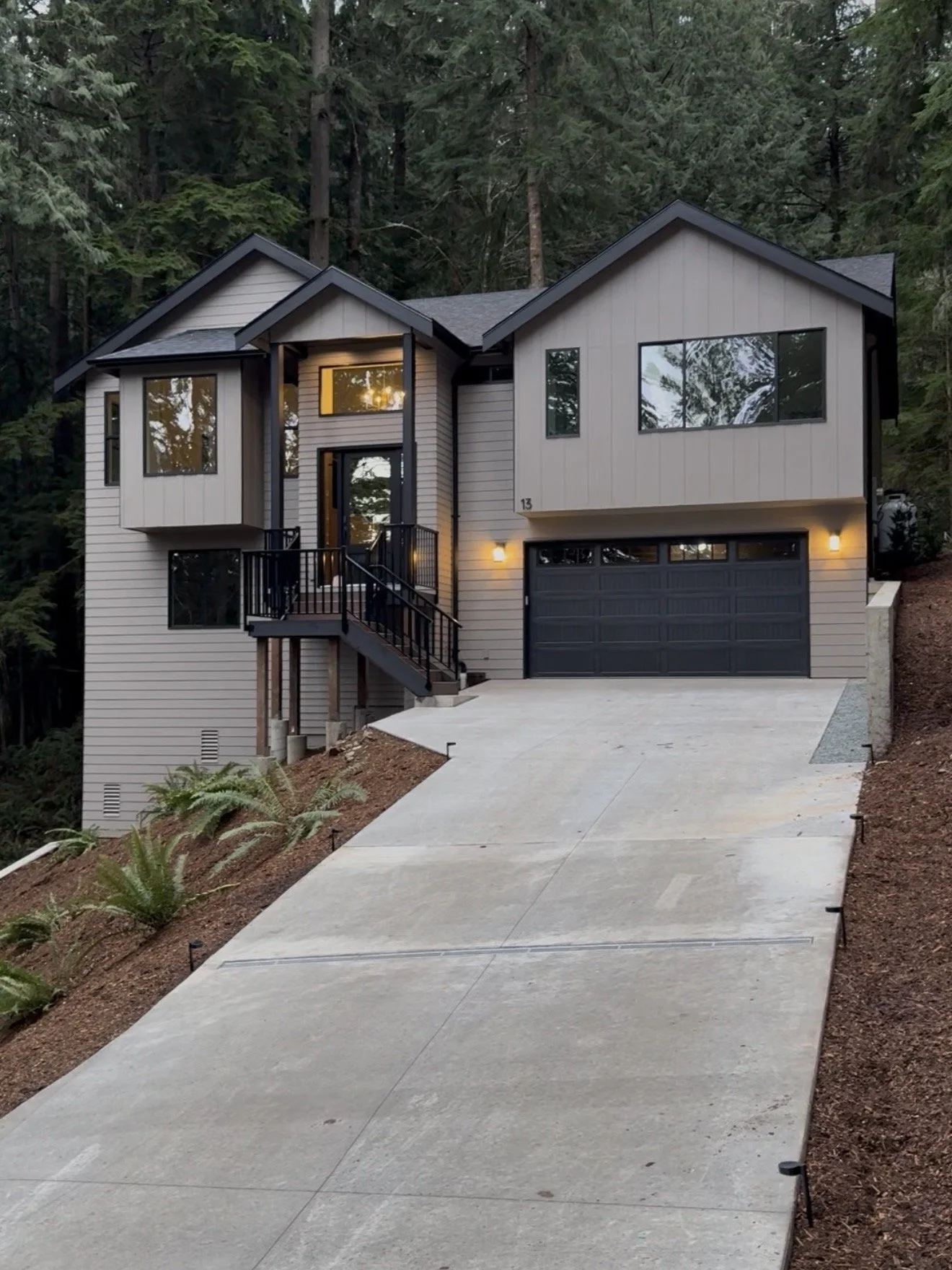 Modern two-story house with gray siding and black accents, surrounded by trees. Features a concrete driveway and a small staircase leading to the front porch, with exterior lights turned on at dusk.
