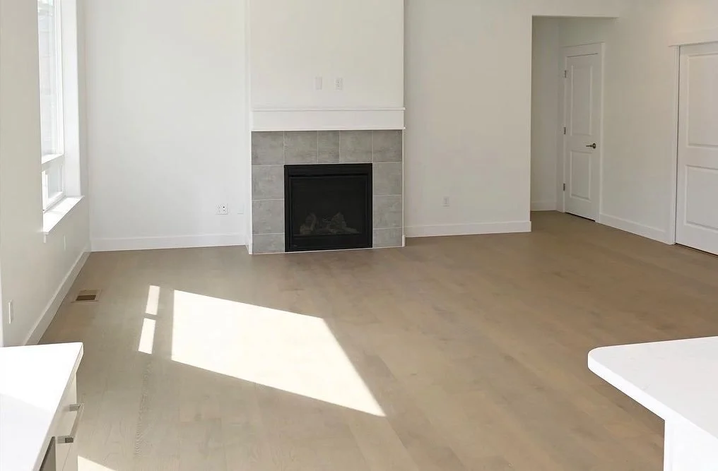 Empty living room with a fireplace, a window with sunlight, white walls, beige hardwood floors, and two white doors.