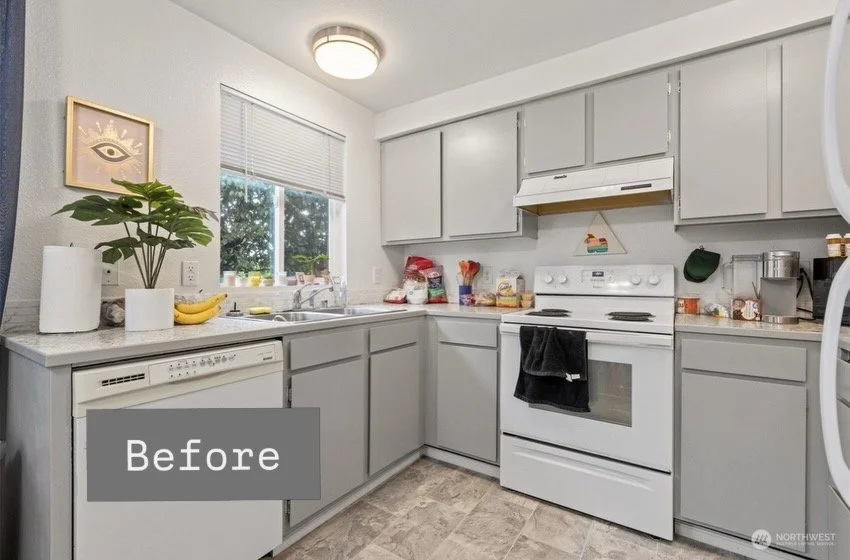 A small kitchen with light gray cabinets, a white stove, and a window with blinds. Countertops have some food items and kitchen appliances, and there is a wall art piece with an eye design on the left.