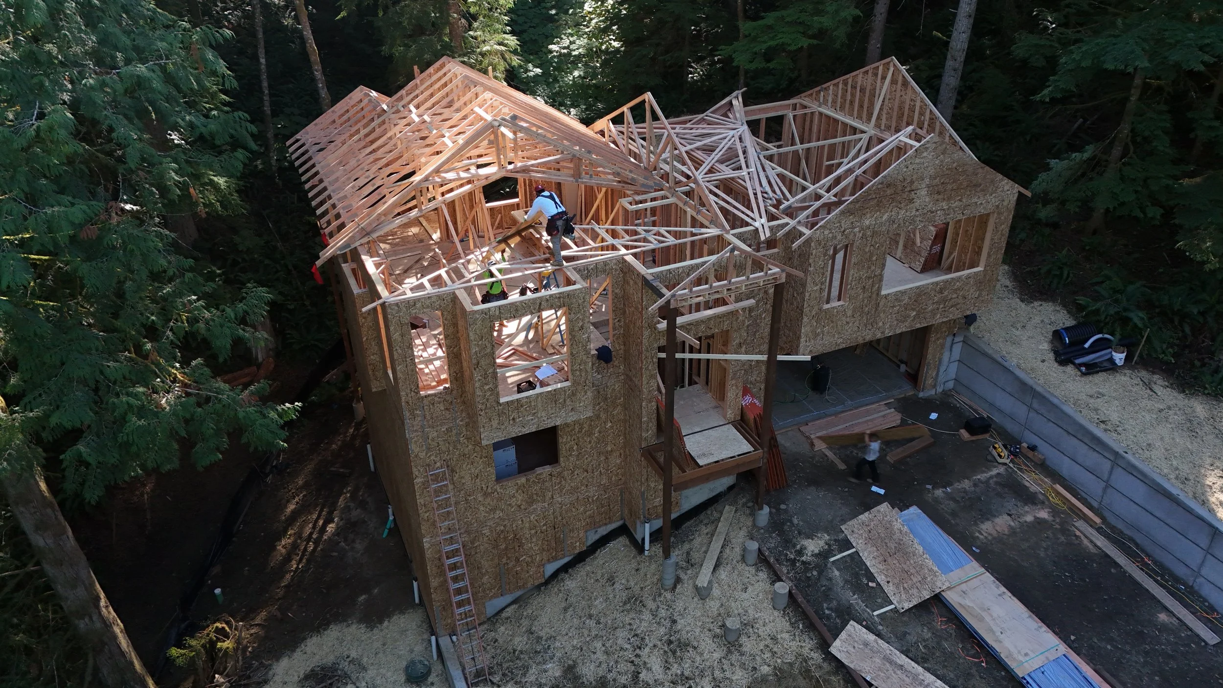 Construction workers building the wooden framework of a multi-story house in a wooded area.