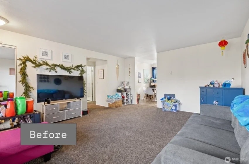 Living room with a large flat-screen TV on a white entertainment unit, decorated with a green garland. To the left, colorful cups on a black table, and a pink chair. On the right, a gray couch and a blue cabinet with decorations. A small hallway and 