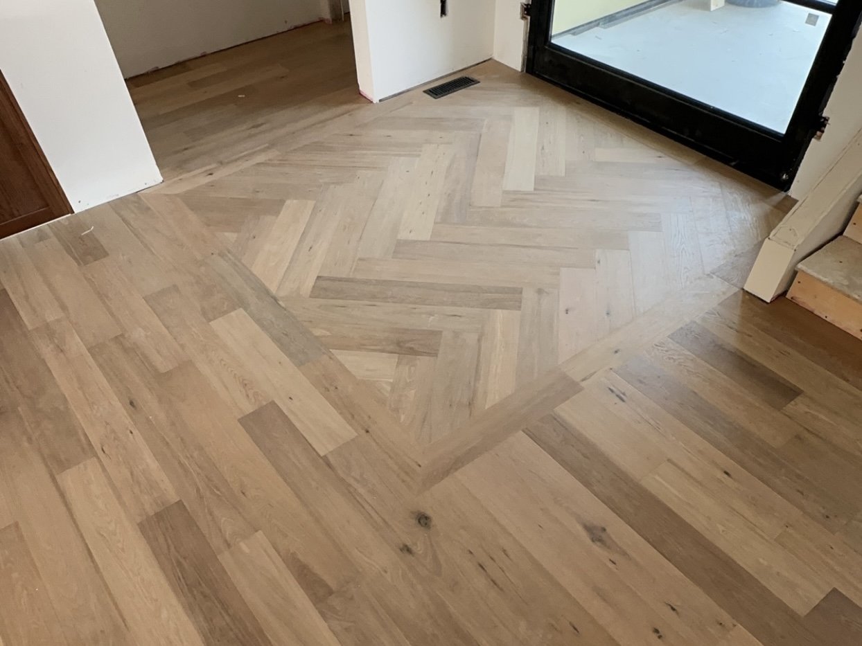 Interior view of a hardwood floor with a herringbone pattern near a glass door, with a mixture of light and dark wood planks, and a vent on the floor.