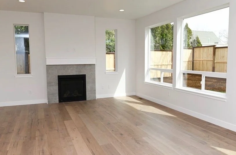Empty living room with hardwood floors, a fireplace with a gray tile surround, and large windows overlooking a backyard with a wooden fence.
