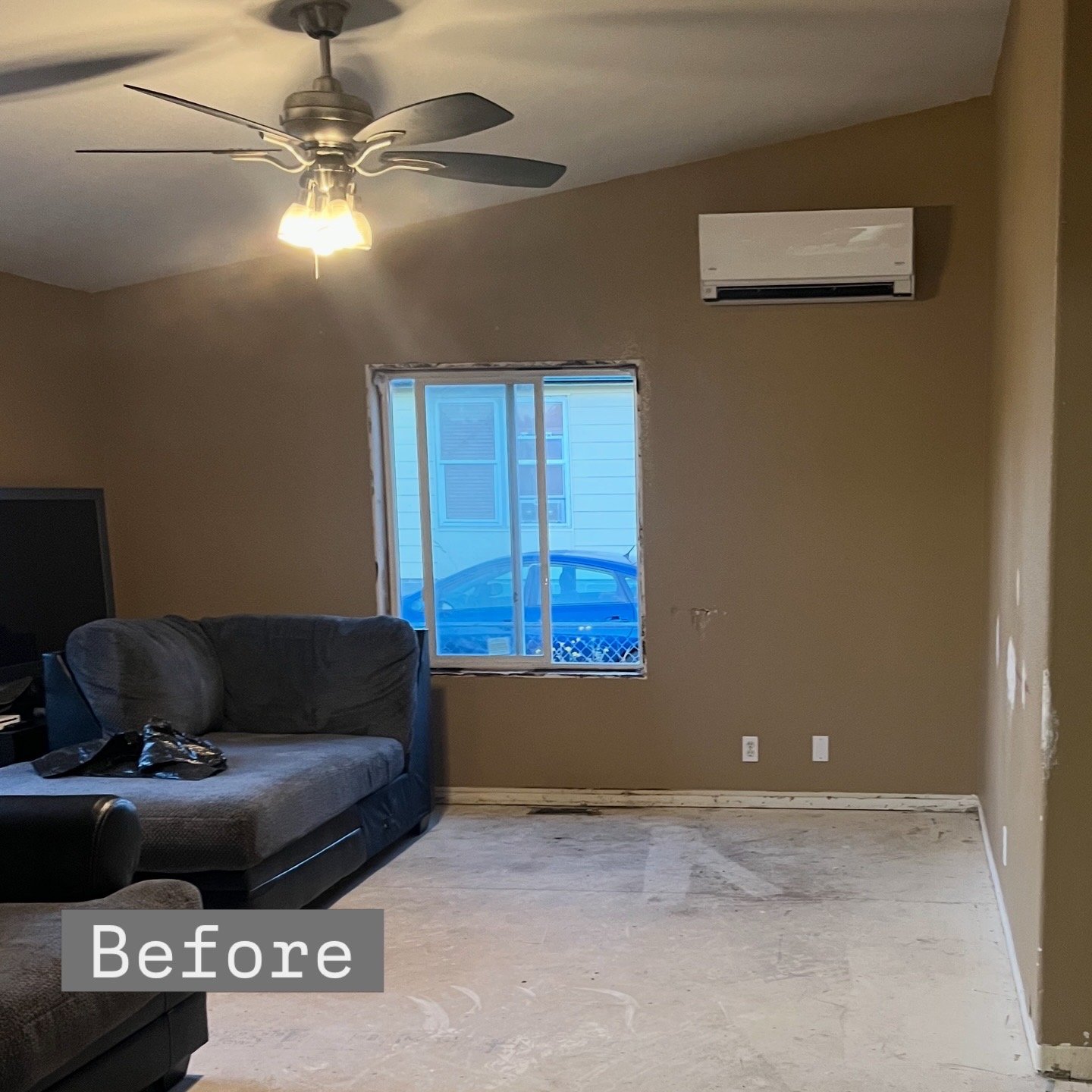 Living room with beige walls, ceiling fan, small window, and air conditioning unit; wall near window has holes and dirt on the floor.