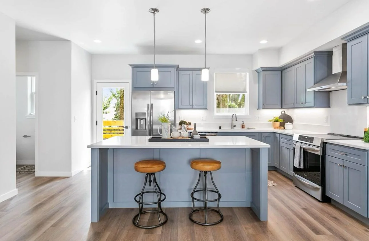 Modern kitchen with blue cabinets, stainless steel refrigerator, stove, and white island with two brown stools, wood flooring, and windows letting in natural light.