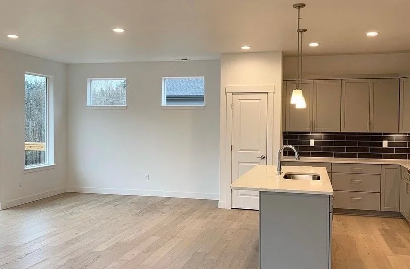 Empty modern kitchen with beige cabinets, black backsplash, small island with sink, open living space, and three small windows, bright and minimalist design.