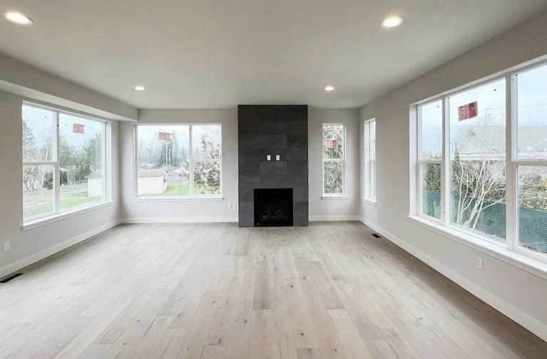 Empty living room with large windows, light wood flooring, and a black fireplace on a dark gray accent wall