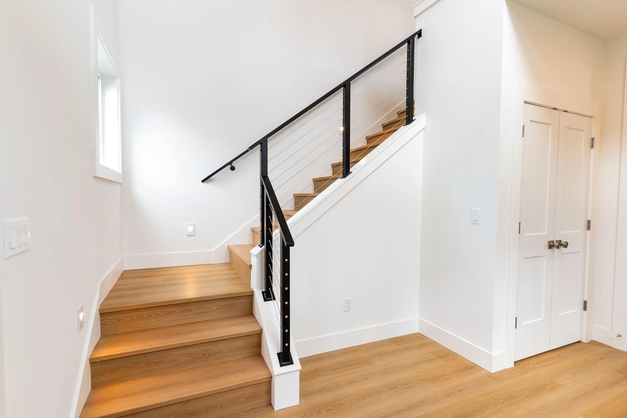Interior view of a staircase with wooden steps, white walls, and a black metal and cable railing, next to white double doors and a small window.