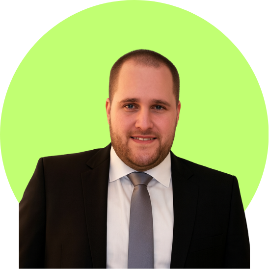 A man with a short beard and mustache, wearing a black suit, white shirt, and gray tie, smiling indoors near a window with blinds.