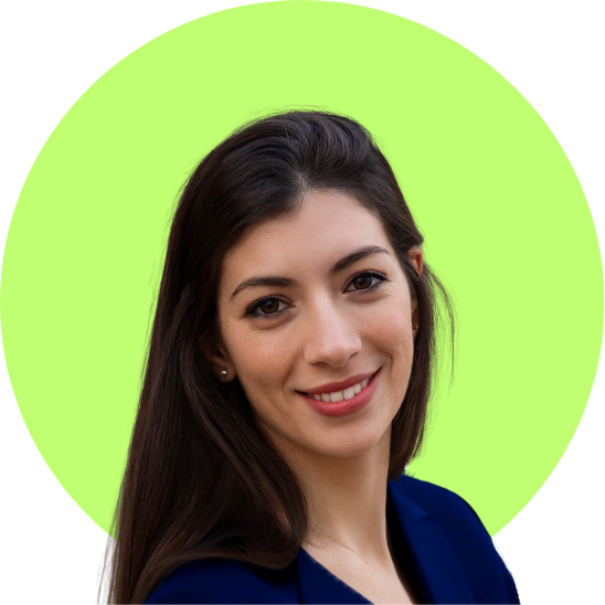 Young woman with long dark hair smiling, wearing a beige sweater, standing against a plain wall.