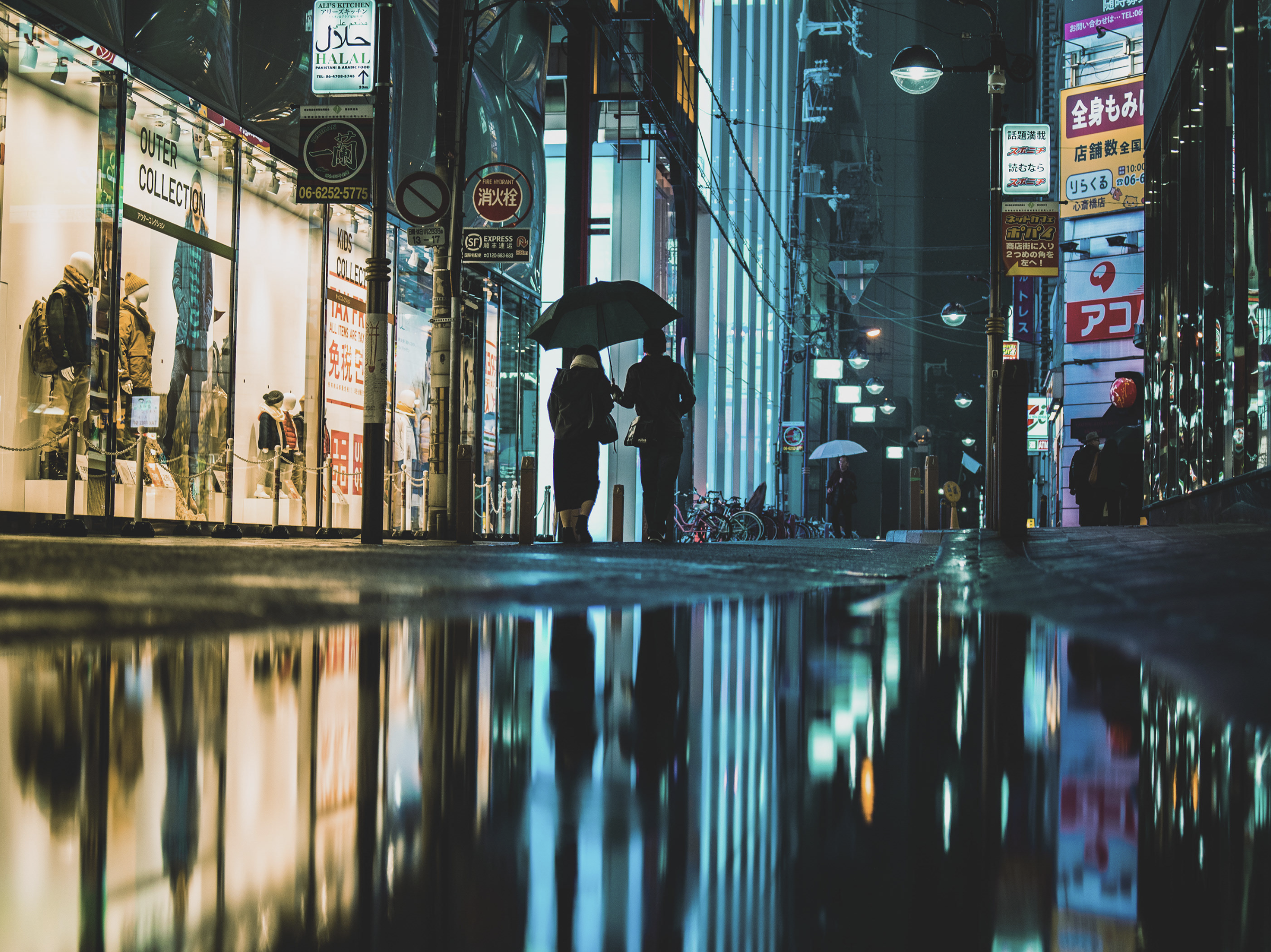 Two people walking under umbrellas in a neon-lit city street at night, reflections of the city lights visible on a wet sidewalk.
