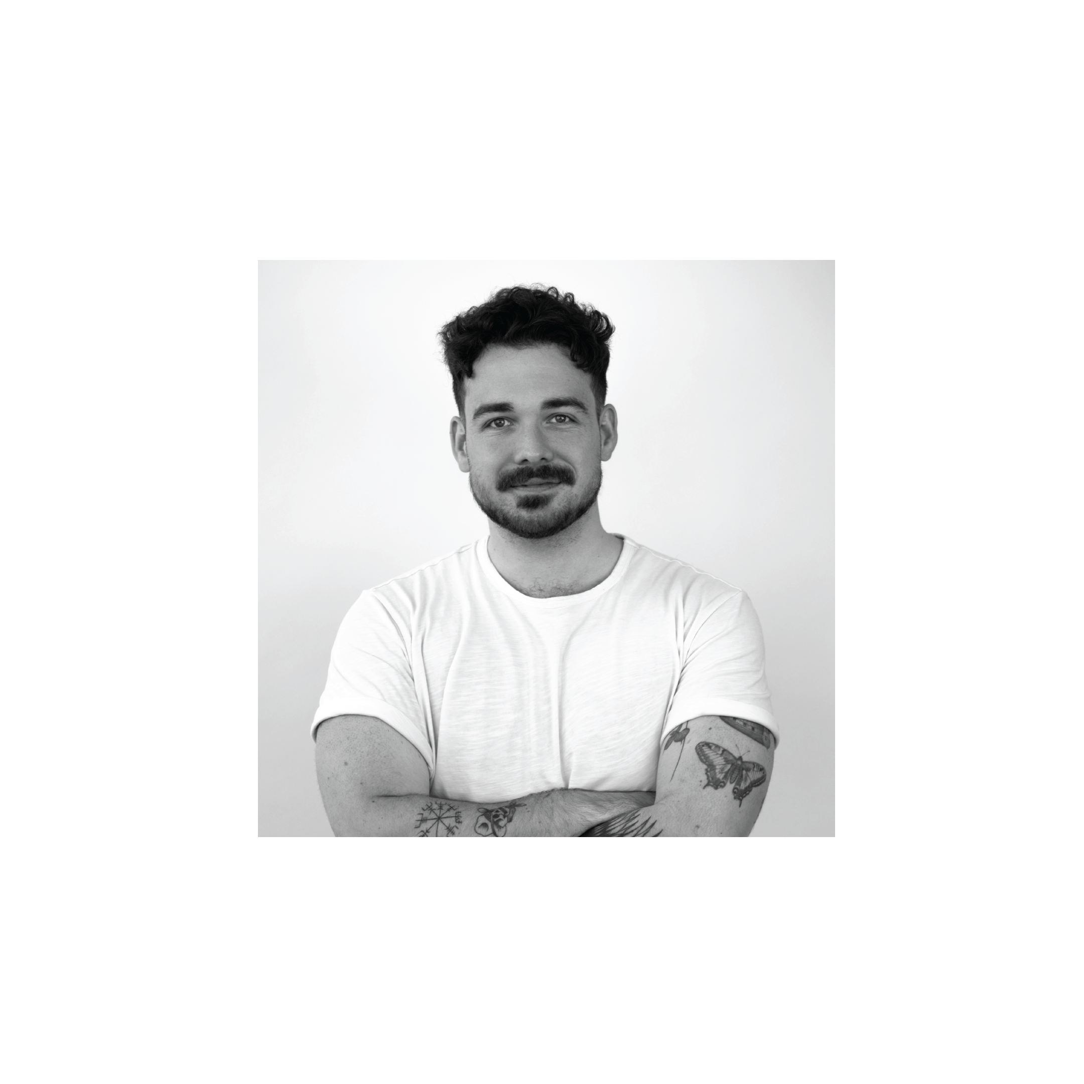 Black and white portrait of a man with short curly hair and a beard, leaning with his arms crossed, wearing a plain white t-shirt, in front of a geometric black and white background.