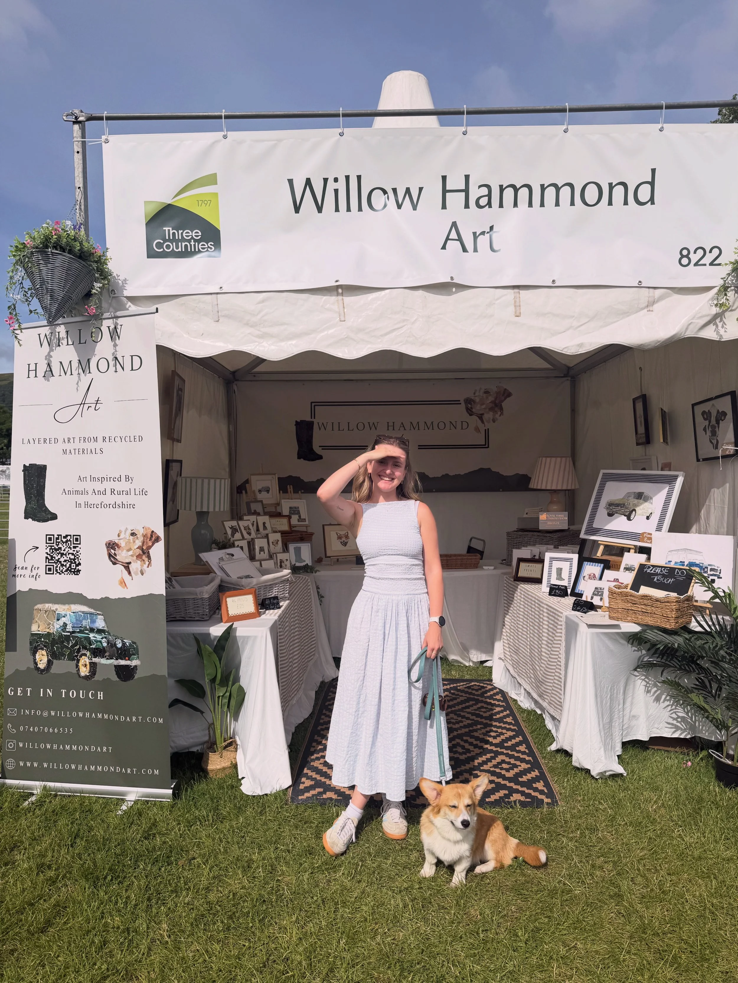 A young woman standing outside a white tent in a grassy area, saluting with her right hand, with a corgi dog sitting on the ground beside her. The tent has a sign and a banner for Willow Hammond Art, showcasing artwork and contact information.