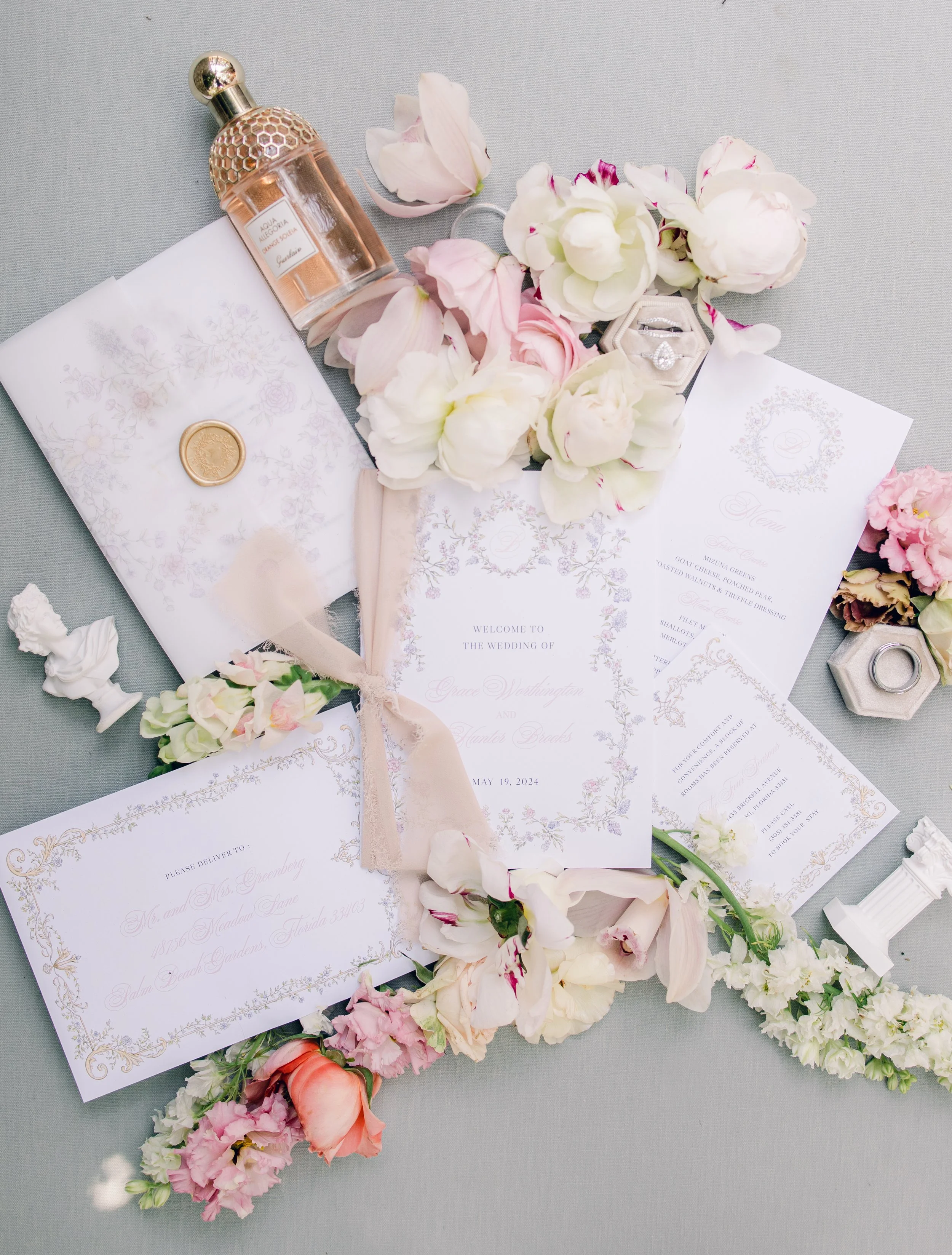 Wedding invitation suite with floral accents, perfume bottle, engagement ring, and wedding rings displayed on a table.