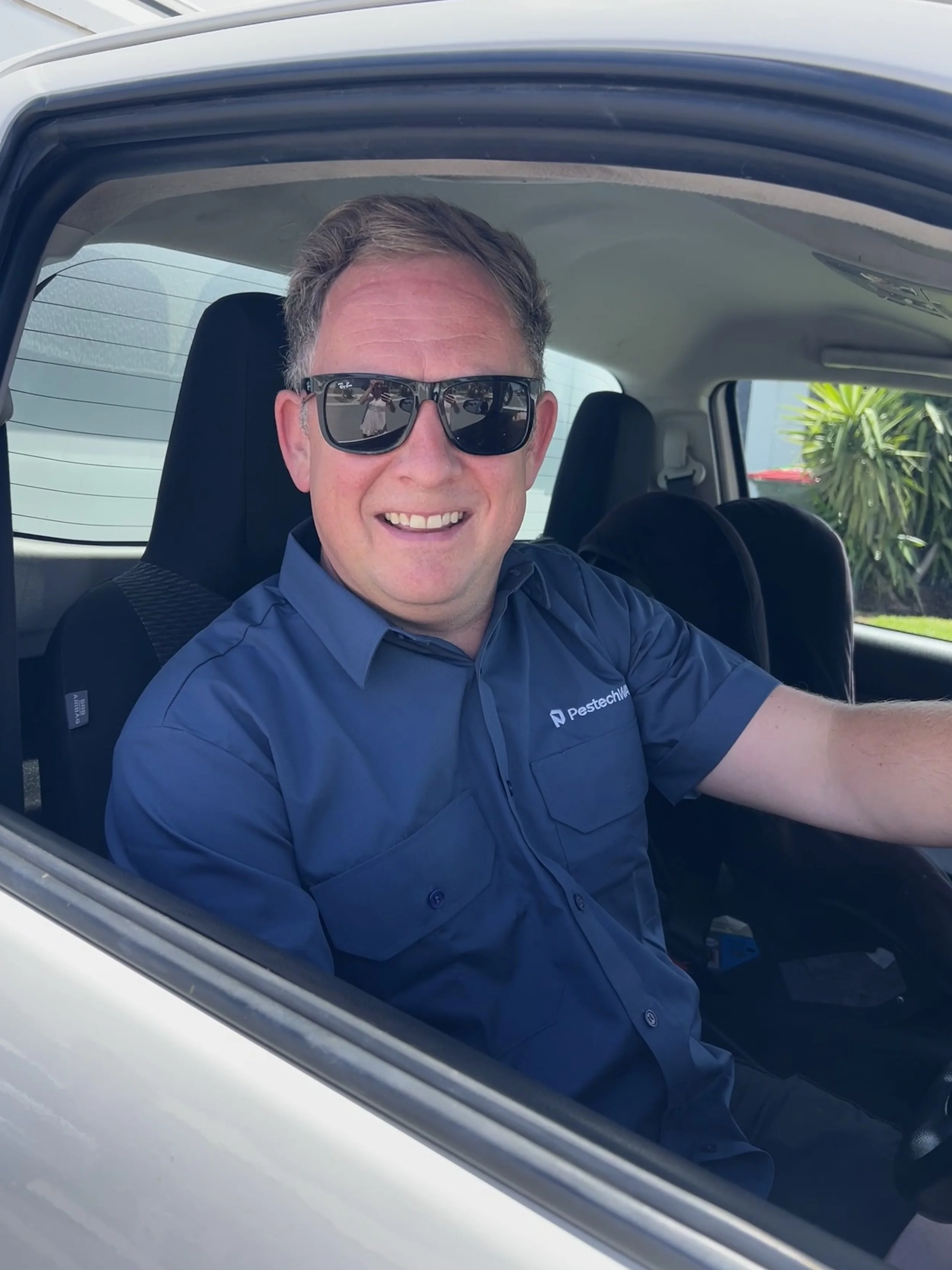 A man smiling and wearing sunglasses sitting inside a vehicle, with a palm tree visible outside the window.