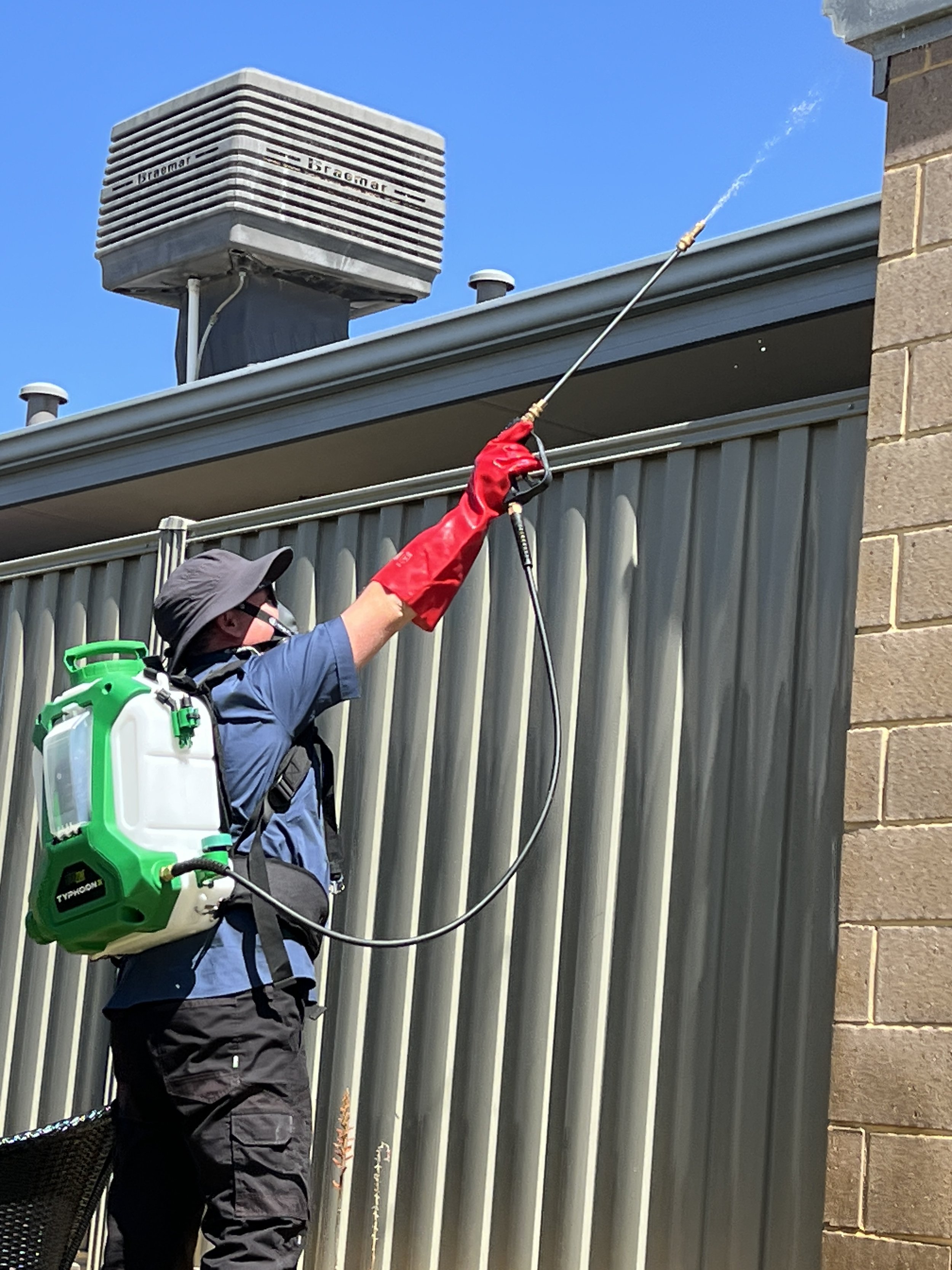 A person wearing a hat, mask, blue shirt, and red gloves using a sprayer to disinfect a metal fence outdoors under a clear blue sky.