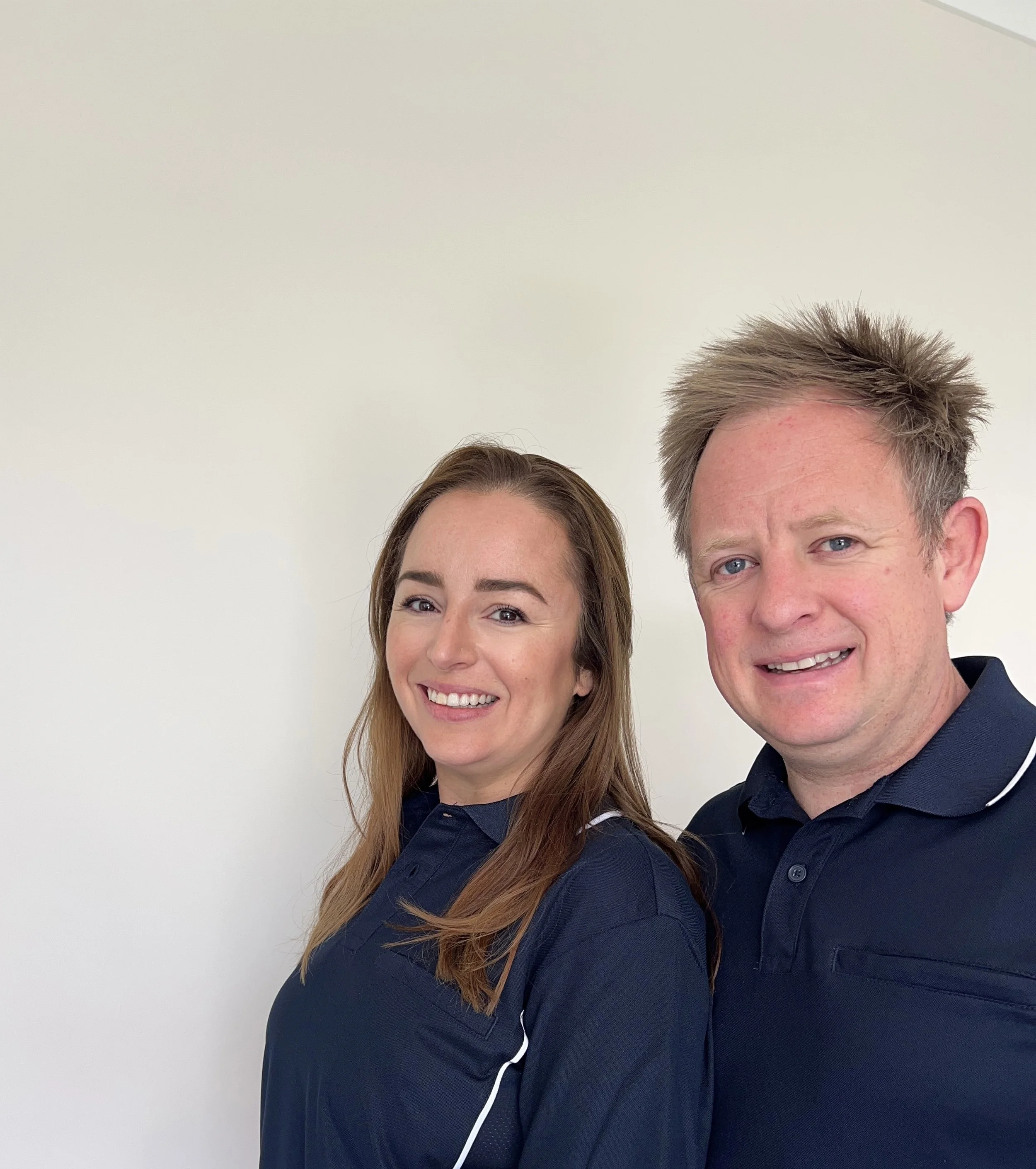 A man and a woman standing side by side, both wearing navy blue collared shirts, smiling against a plain light-colored background.