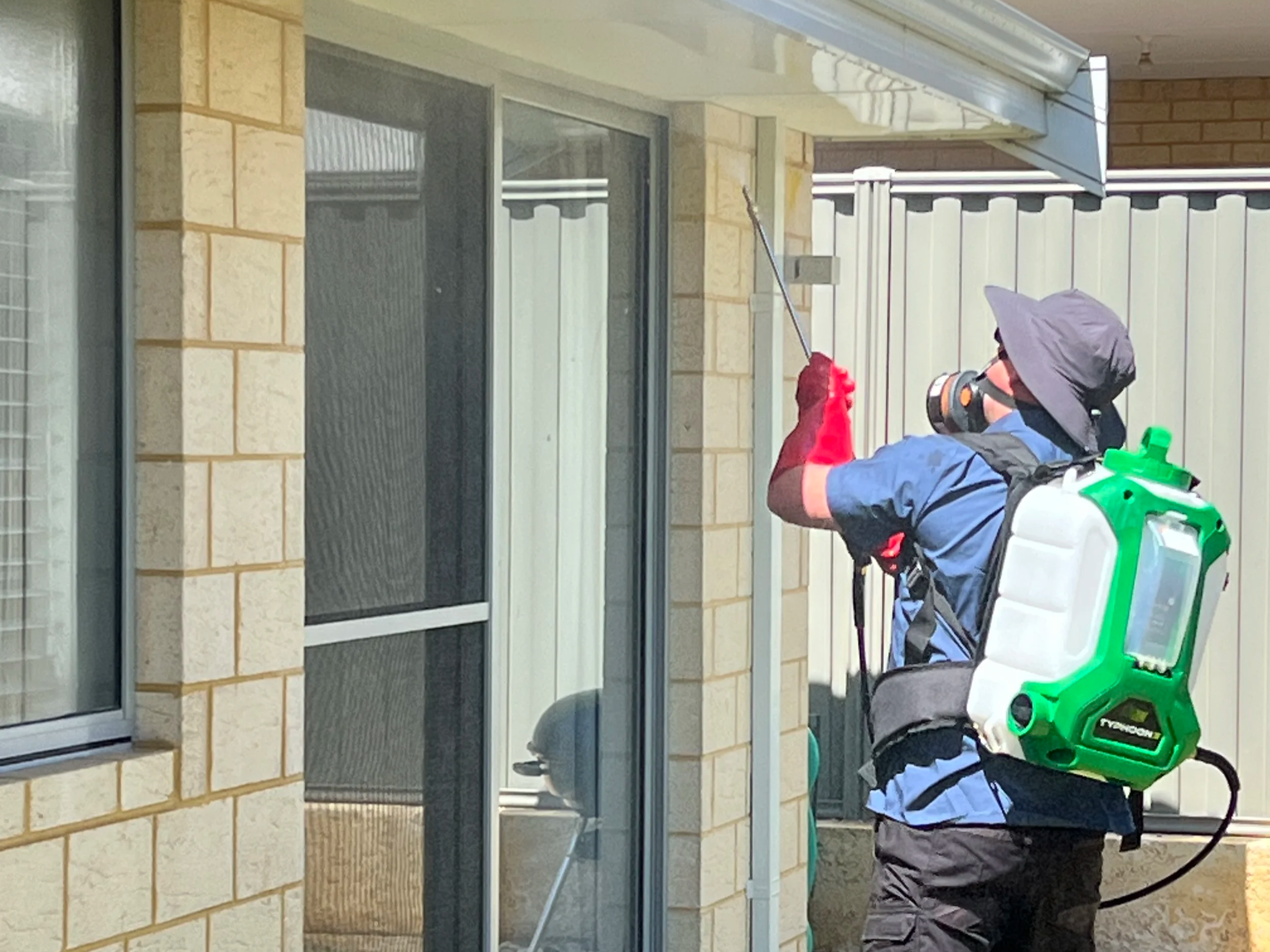 A person wearing a wide-brim hat, mask, gloves, and a backpack sprayer is disinfecting or spraying the exterior of a building through a window using a long spray wand.