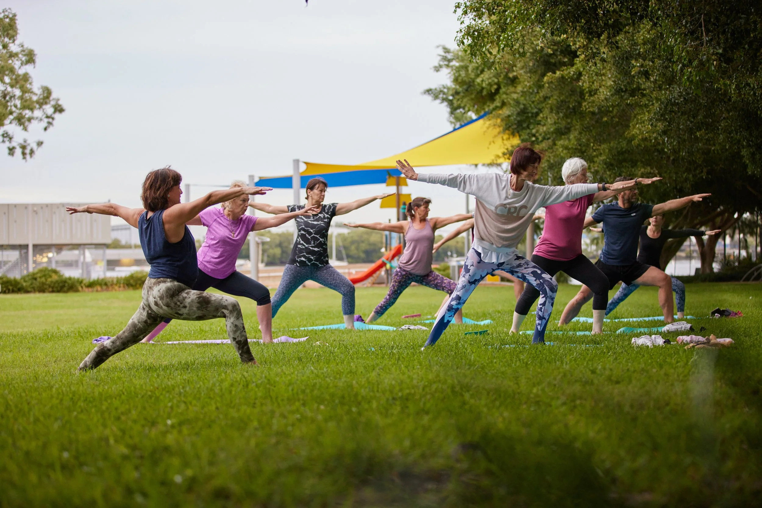 Group of women practicing yoga outdoors on a grassy field, performing warrior pose with arms extended and one leg forward.