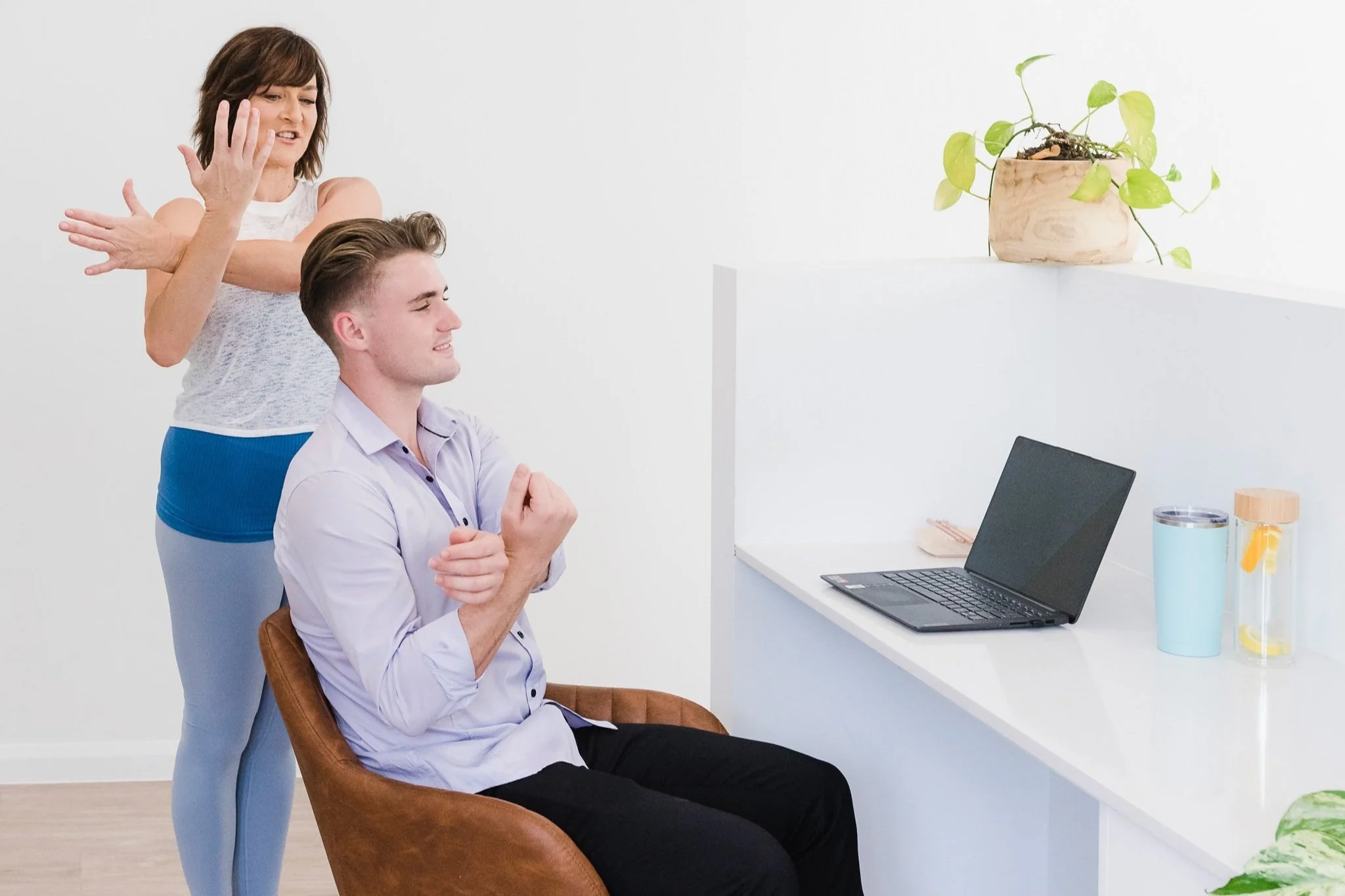 A man sitting in a chair with his fists clenched, smiling, in front of a laptop. A woman behind him appears to be stretching or practicing yoga.
