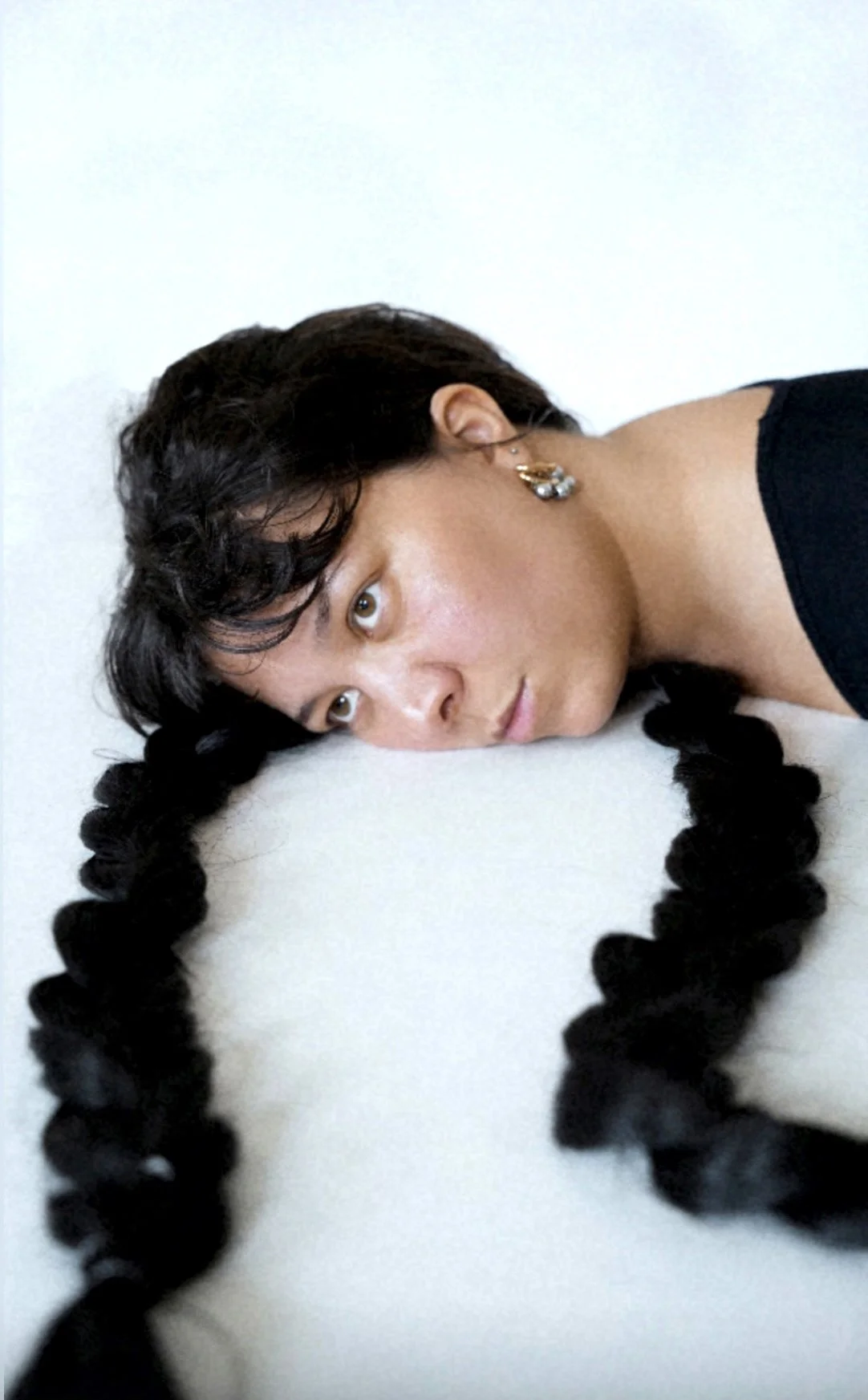 Femme avec deux longues tresses noires, allongée sur le sol avec la tête reposant sur le côté, regardant fixement en direction de l'appareil, portant une boucle d'oreille dorée et noire, vêtue d'un vêtement noir.