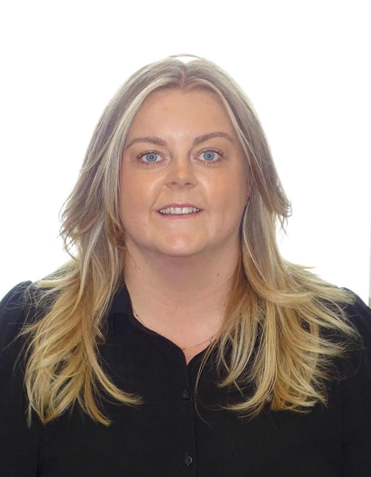 A woman with blonde, wavy hair and blue eyes, wearing a black dress, smiling at the camera against a white background.